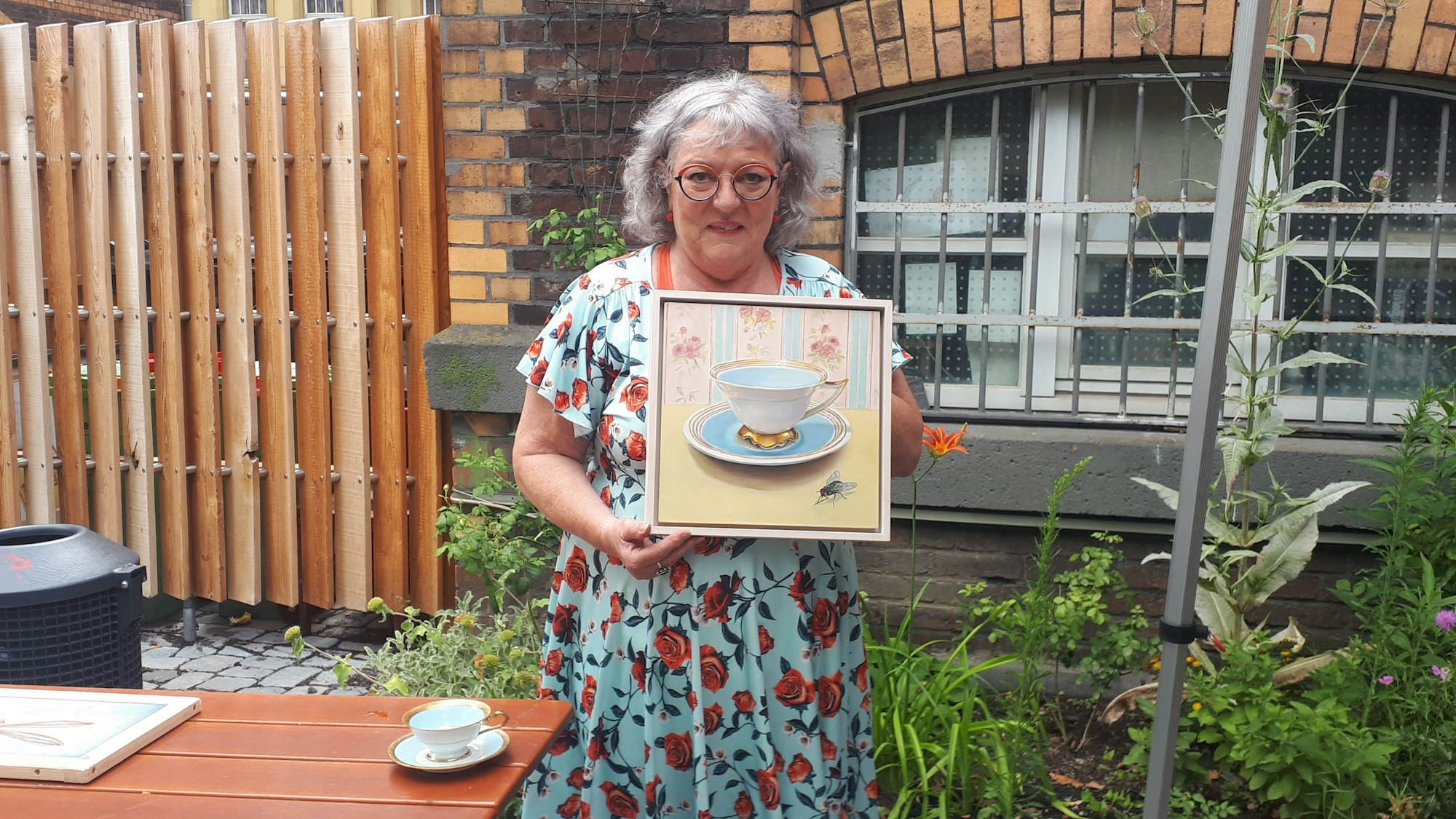 Eine Frau mit grauen Haaren trägt ein hellblaues Kleid mit roten Rosen darauf. Sie steht in einem Garten und hält ein Bild, auf dem eine historische Tasse und eine Fliege zu erkennen sind.