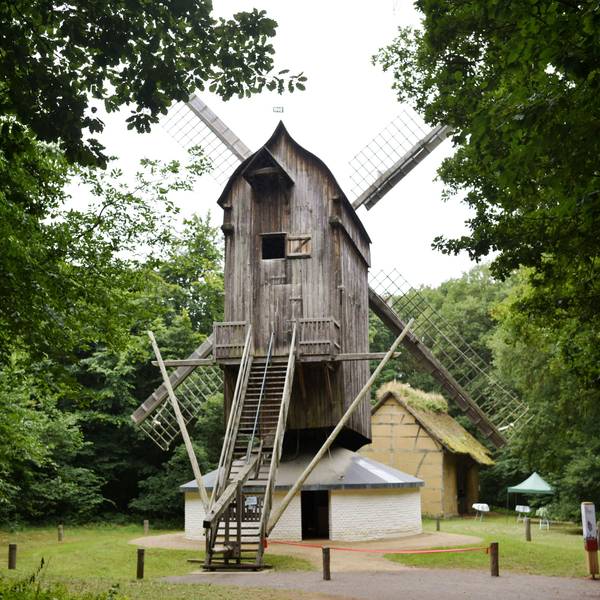 Eine Bockwindmühle aus Holz steht zwischen grünen Bäumen.