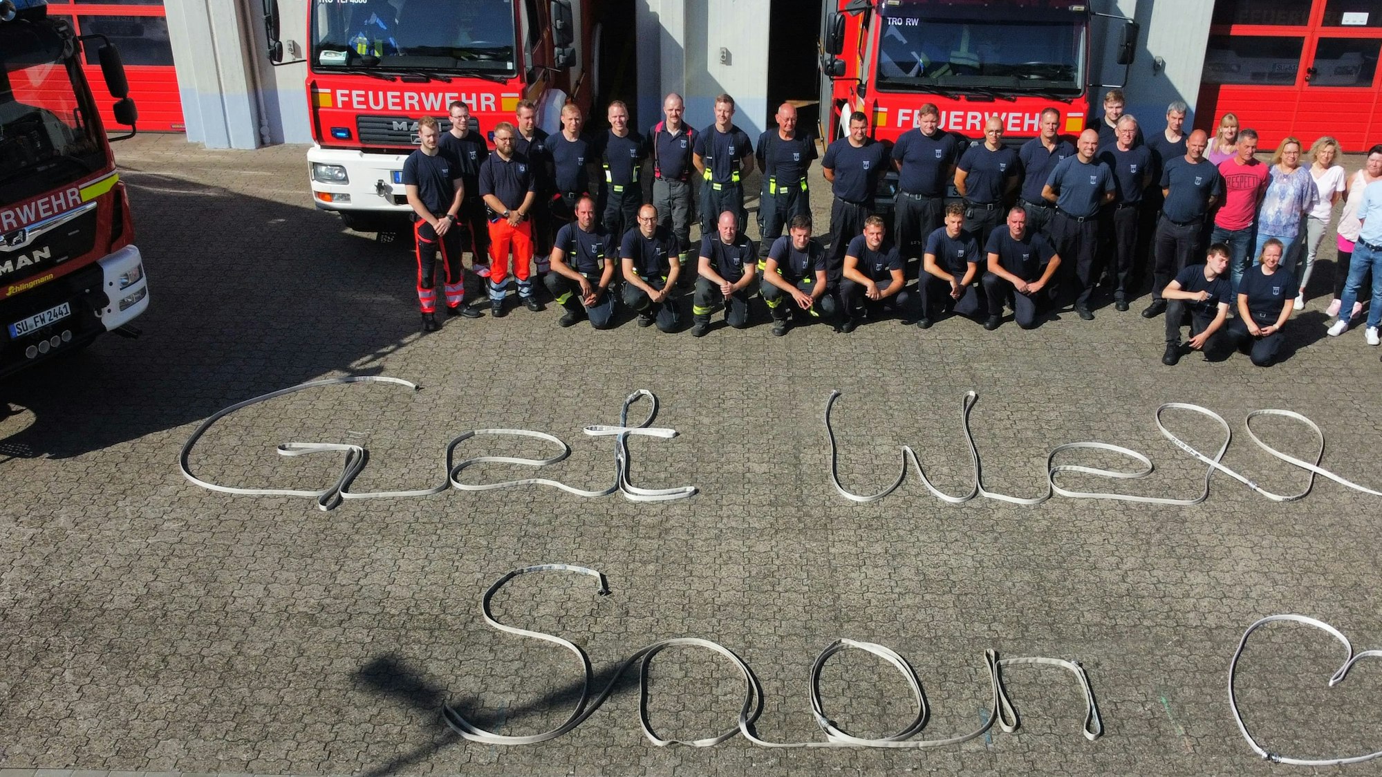 Feuerwehrleute vor einer Fahrzeughalle; auf dem Boden ist der Satz „Get well soon“ zu lesen.