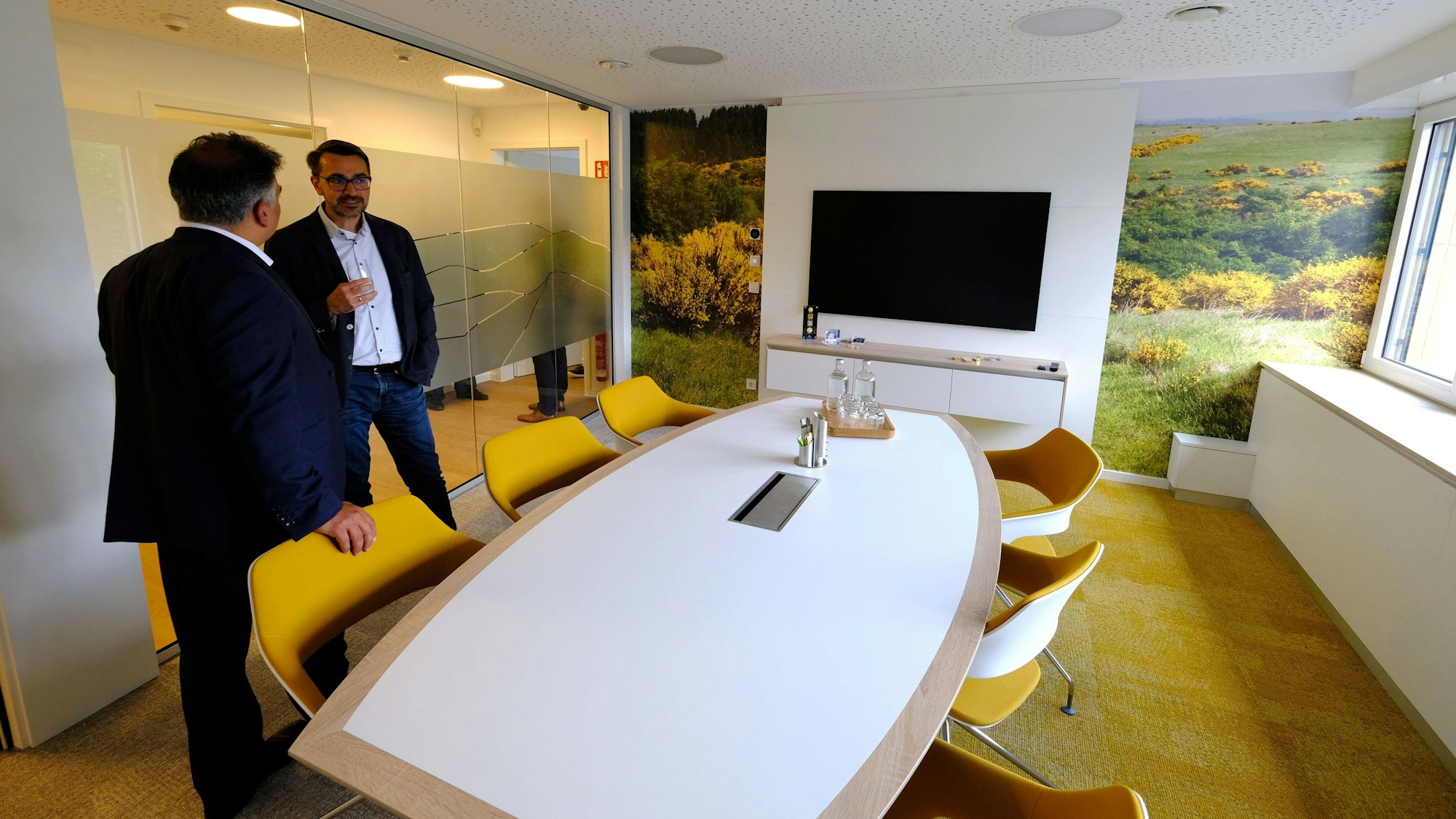 Zwei Männer stehen in einem modern gestalteten Besprechungsraum, die Wand wird mit Fotos von blühendem Ginster geschmückt.