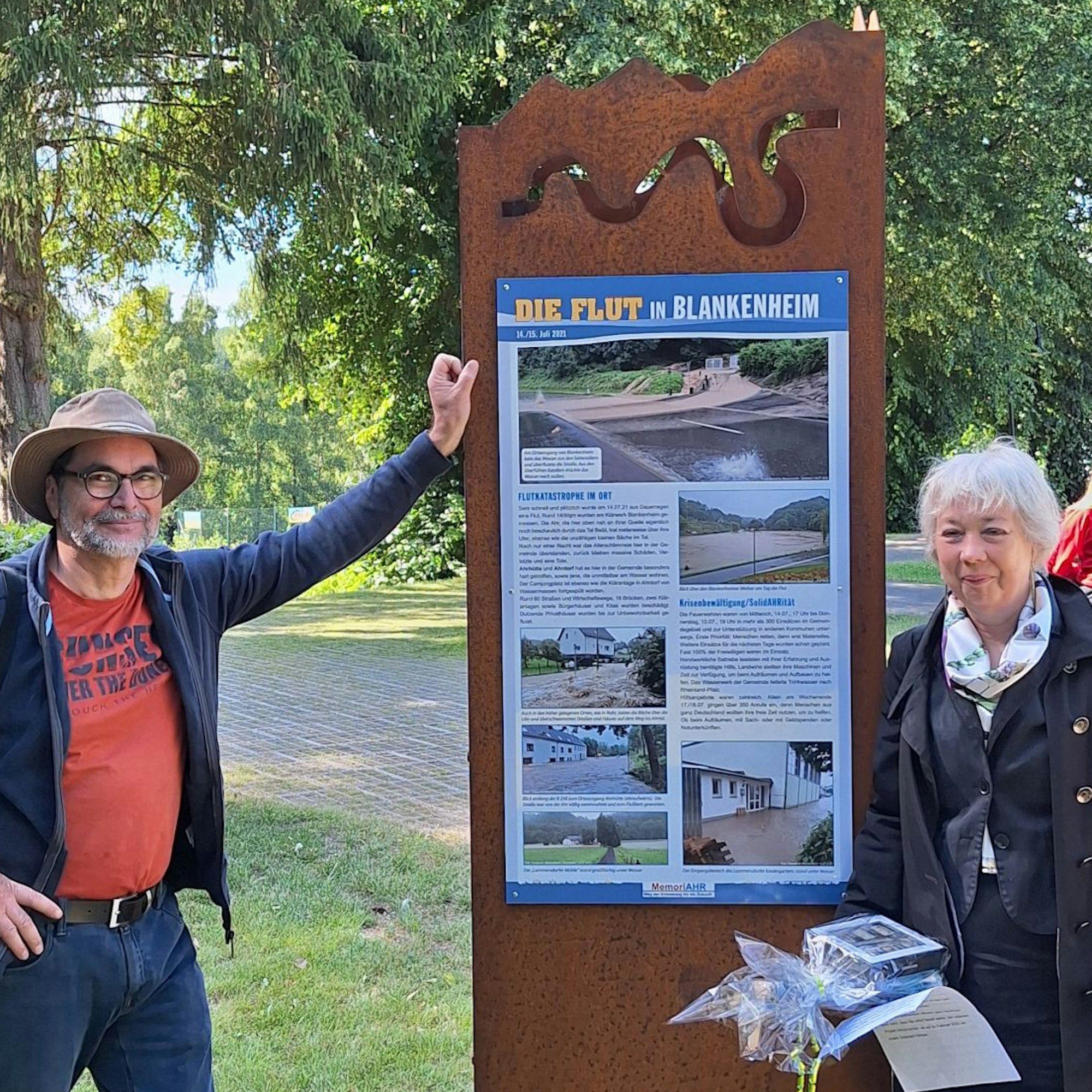 Annette Holzapfel und Andreas Schmickler stehen an einer Stele, auf der „Die Flut in Blankenheim“ steht. Es sind mehrere Bilder zur Flutkatastrophe mit Textelementen versehen.