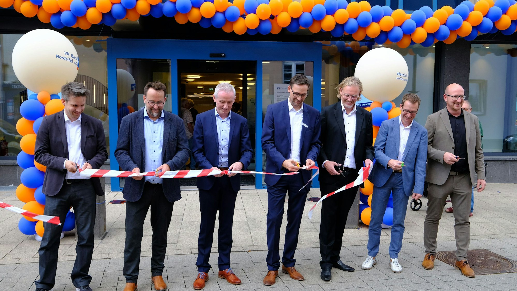 Wiedereröffnung der VR-Bank in Schleiden: Sieben Männer in Anzügen zerschneiden symbolisch ein rot-weißes Flatterband.
