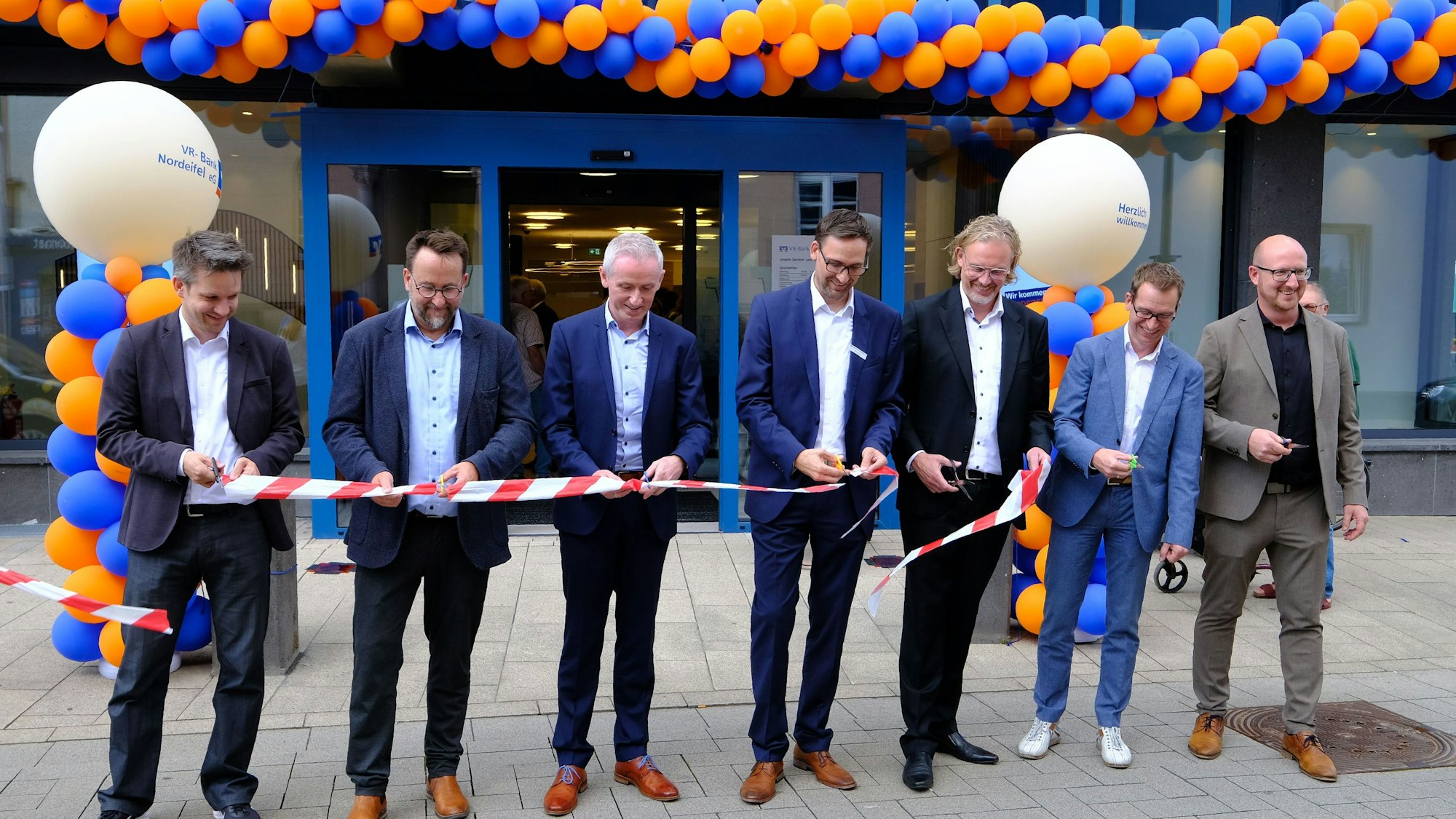 Wiedereröffnung der VR-Bank in Schleiden: Sieben Männer in Anzügen zerschneiden symbolisch ein rot-weißes Flatterband.