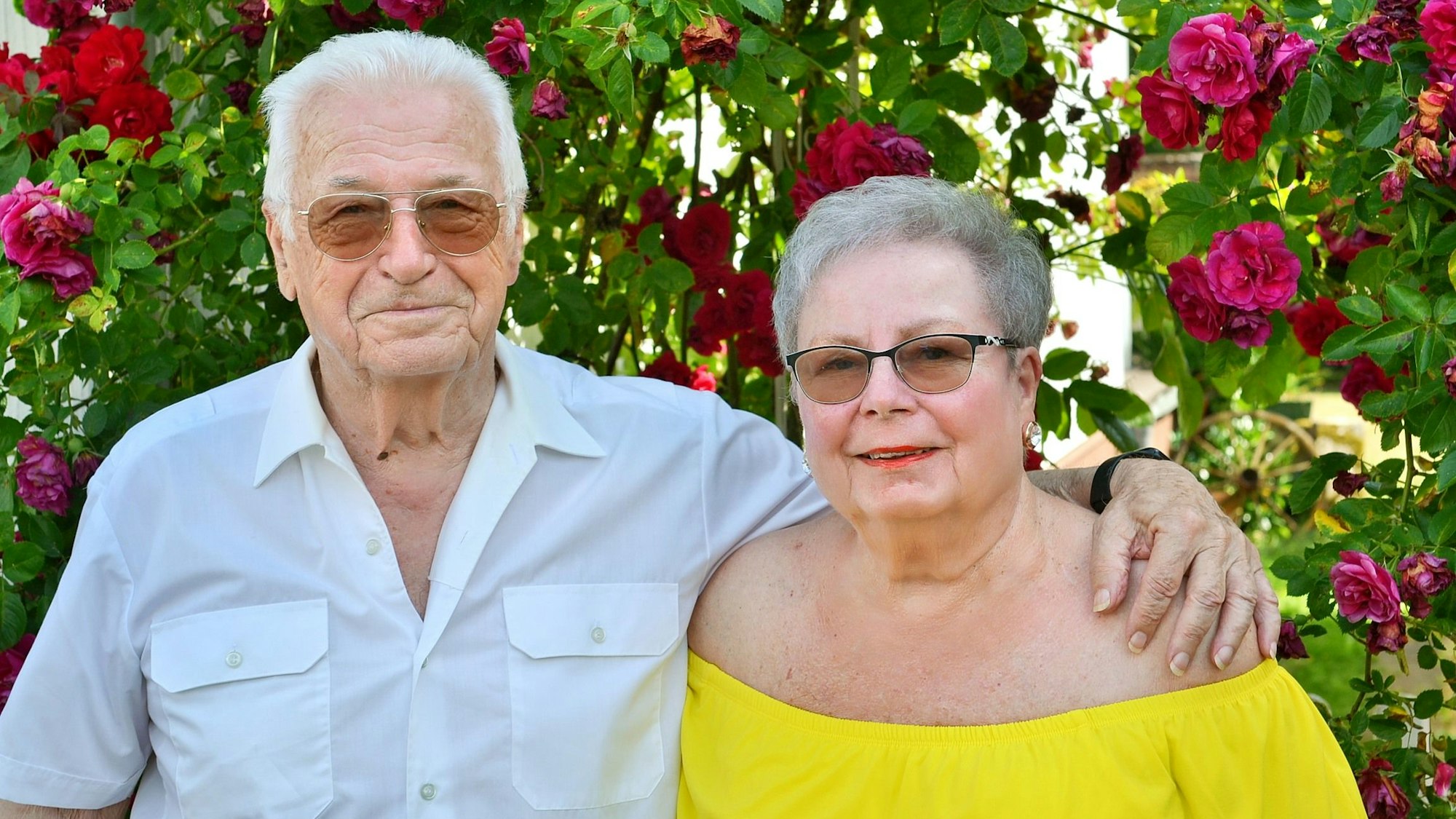 Helmut und Edith Tschernay tragen Sonnenbrillen und stehen eng beisammen in einem Rasen. Im Hintergrund blühen Rosen.