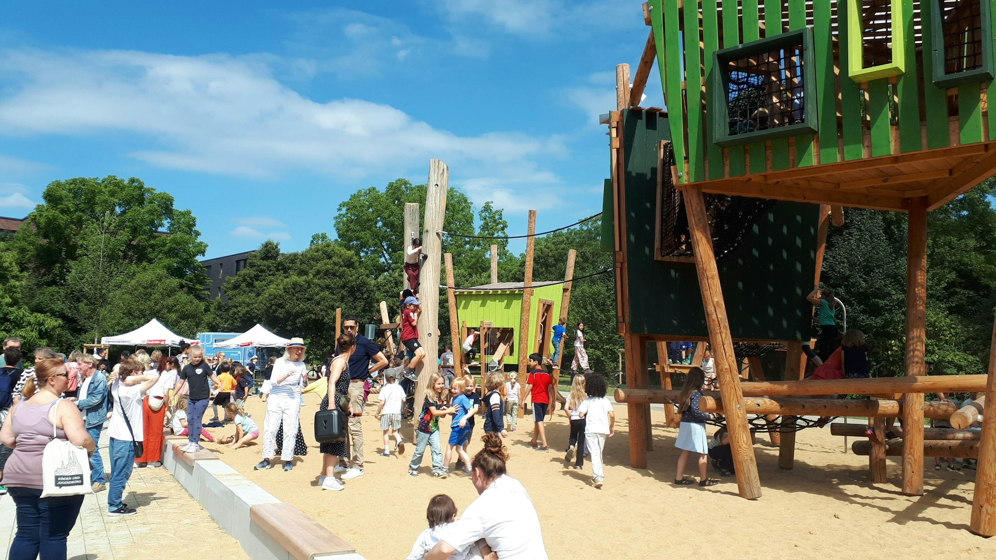 Viele Kinder und Erwachsene sind auf einem neuen Spielplatz mit neuen großen Holz-Klettergeräten.