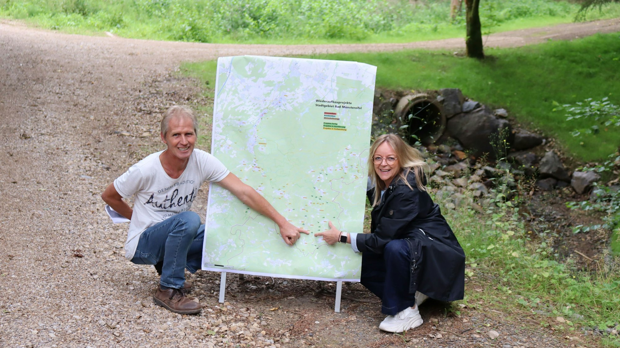 Bernd Hiltrop und Sabine Preiser-Marian knien auf einem Wirtschaftsweg an einer Karte, auf der alle von der Flut beschädigten Wirtschaftswege in der Stadt Bad Münstereifel dargestellt sind.