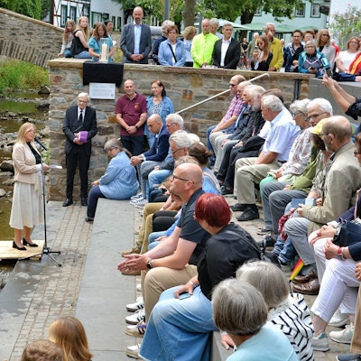 Ein Strauß Sonnenblumen steht in Bad Münstereifel in der Erft. Bürgermeisterin Sabine Preiser-Marian steht auf einem kleinen Podest und spricht in ein Mikrofon. Zahlreiche Menschen sitzen auf Steinstufen und hören ihr zu.
