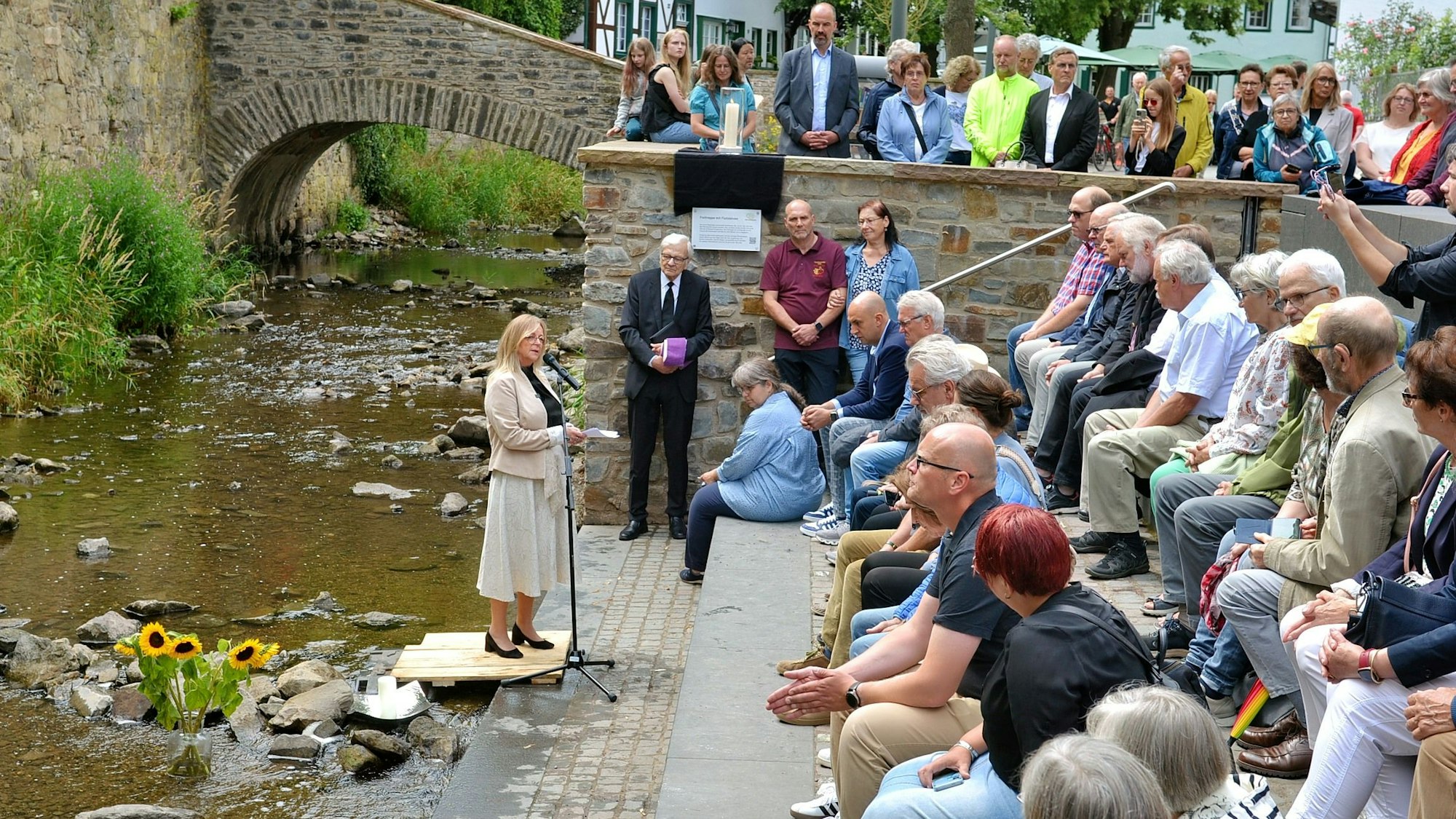 Ein Strauß Sonnenblumen steht in Bad Münstereifel in der Erft. Bürgermeisterin Sabine Preiser-Marian steht auf einem kleinen Podest und spricht in ein Mikrofon. Zahlreiche Menschen sitzen auf Steinstufen und hören ihr zu.