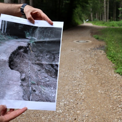 Zwei Hände halten ein Foto, auf dem ein von der Flut zerstörter Wirtschaftsweg in der Stadt Bad Münstereifel zu sehen ist. Rechts im Bild ist der wiederaufgebaute Weg.
