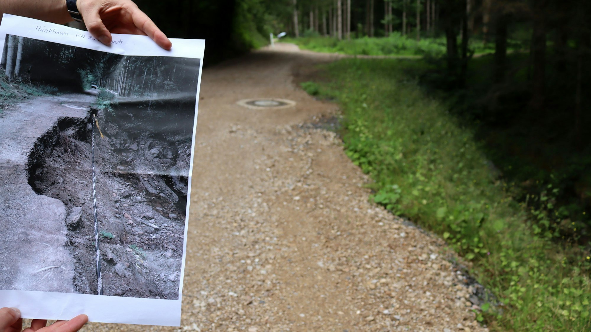Zwei Hände halten ein Foto, auf dem ein von der Flut zerstörter Wirtschaftsweg in der Stadt Bad Münstereifel zu sehen ist. Rechts im Bild ist der wiederaufgebaute Weg.