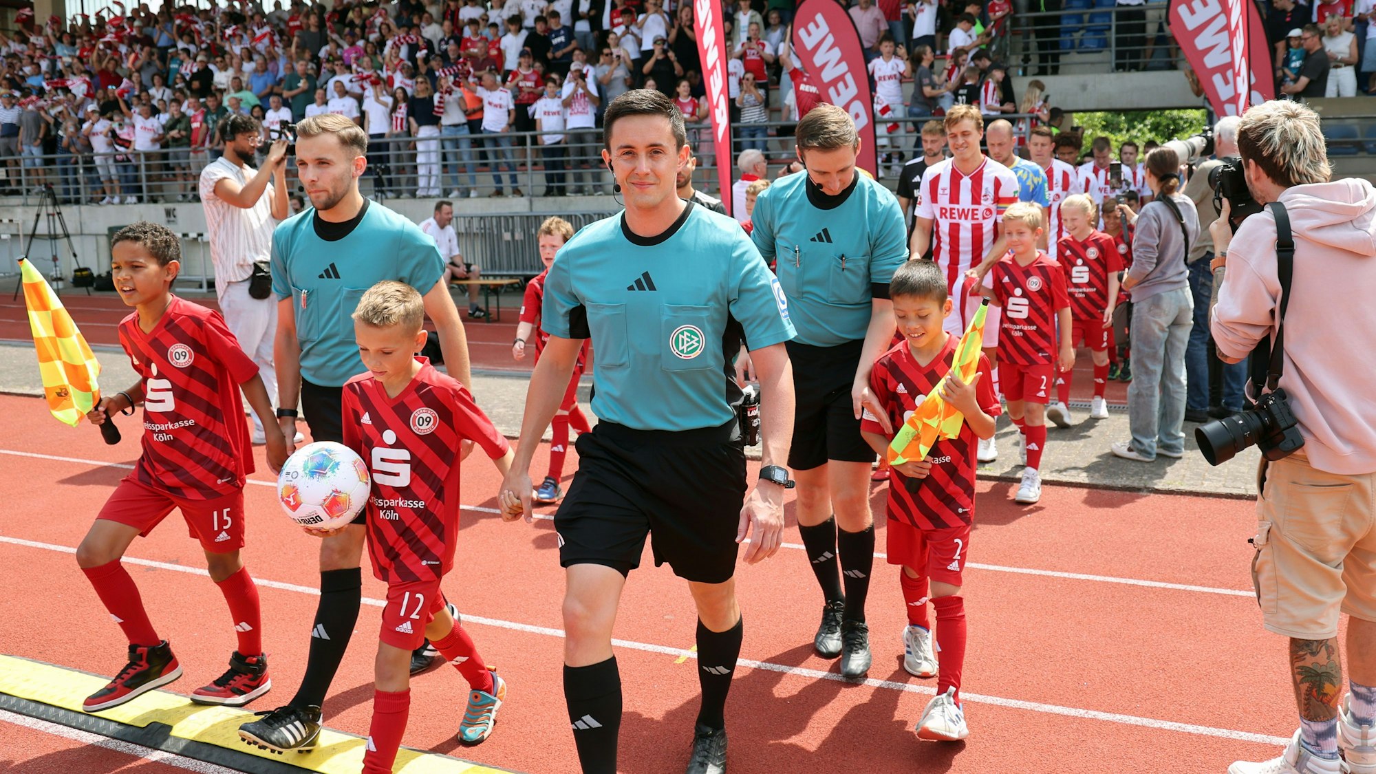 Schiedsrichter mit Nachwuchsspielern an der Hand laufen in ein Fußballstadion ein.