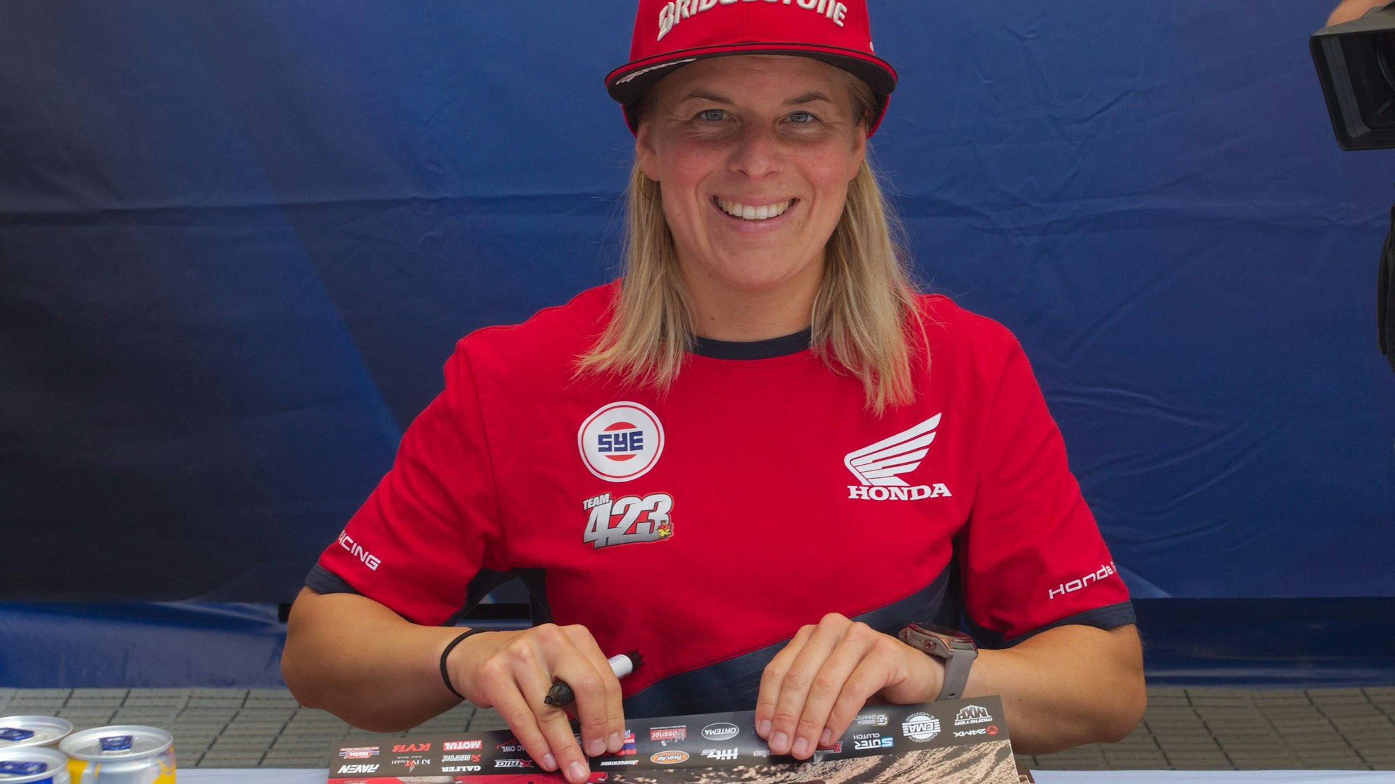Eine Motocrossfahrerin sitzt an einem Tisch und schreibt Autogramme.