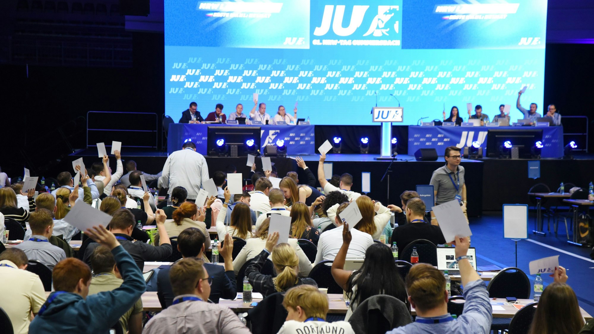 Junge Leute heben Stimmzettel vor einer beleuchteten Podiumsbühne.