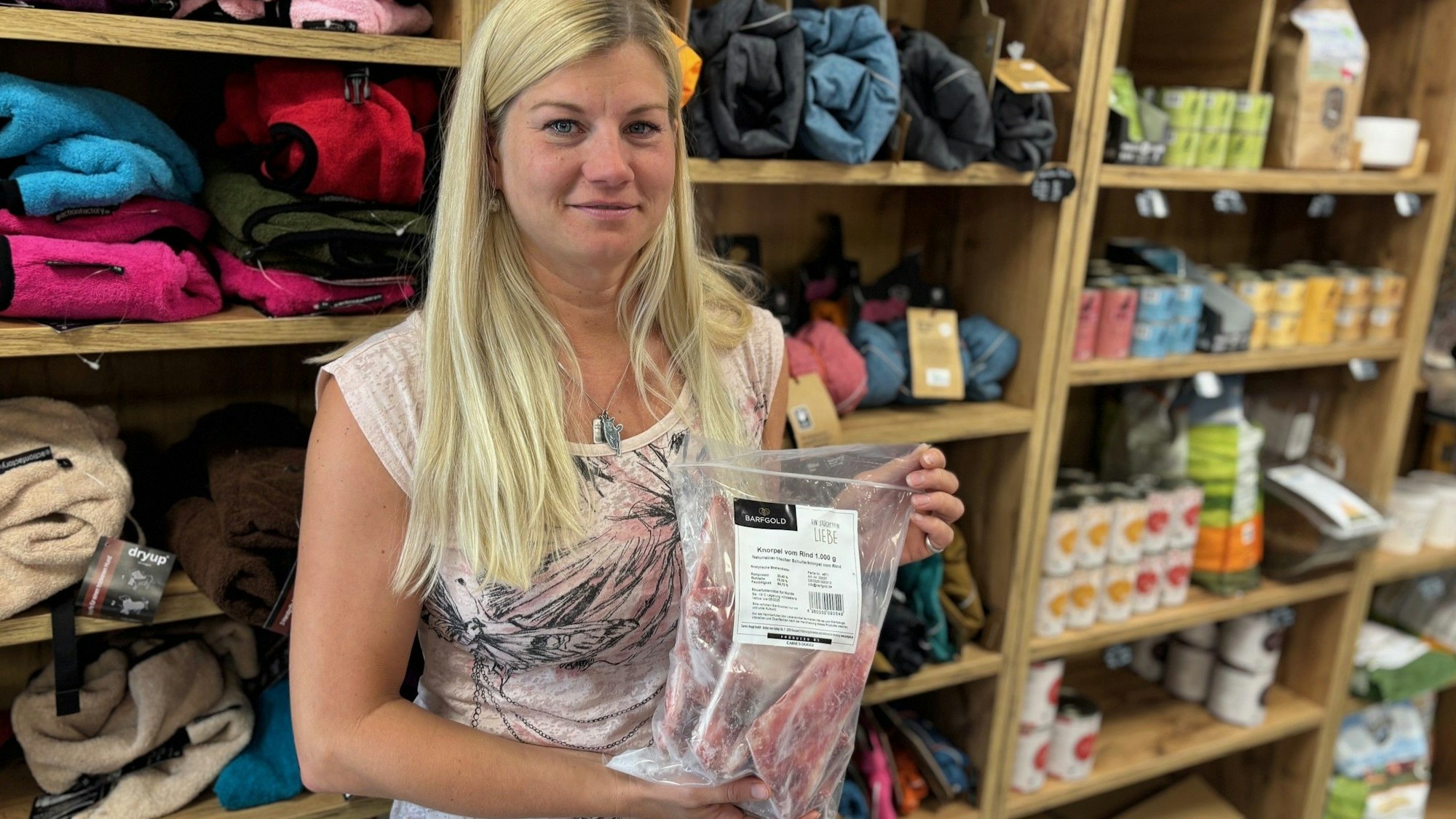 Auf dem Bild ist eine blonde Frau mit einer Tüte Tiefkühlhundefutter vor Holzregalen mit Decken und Pflegemitteln für Hunde zu sehen.