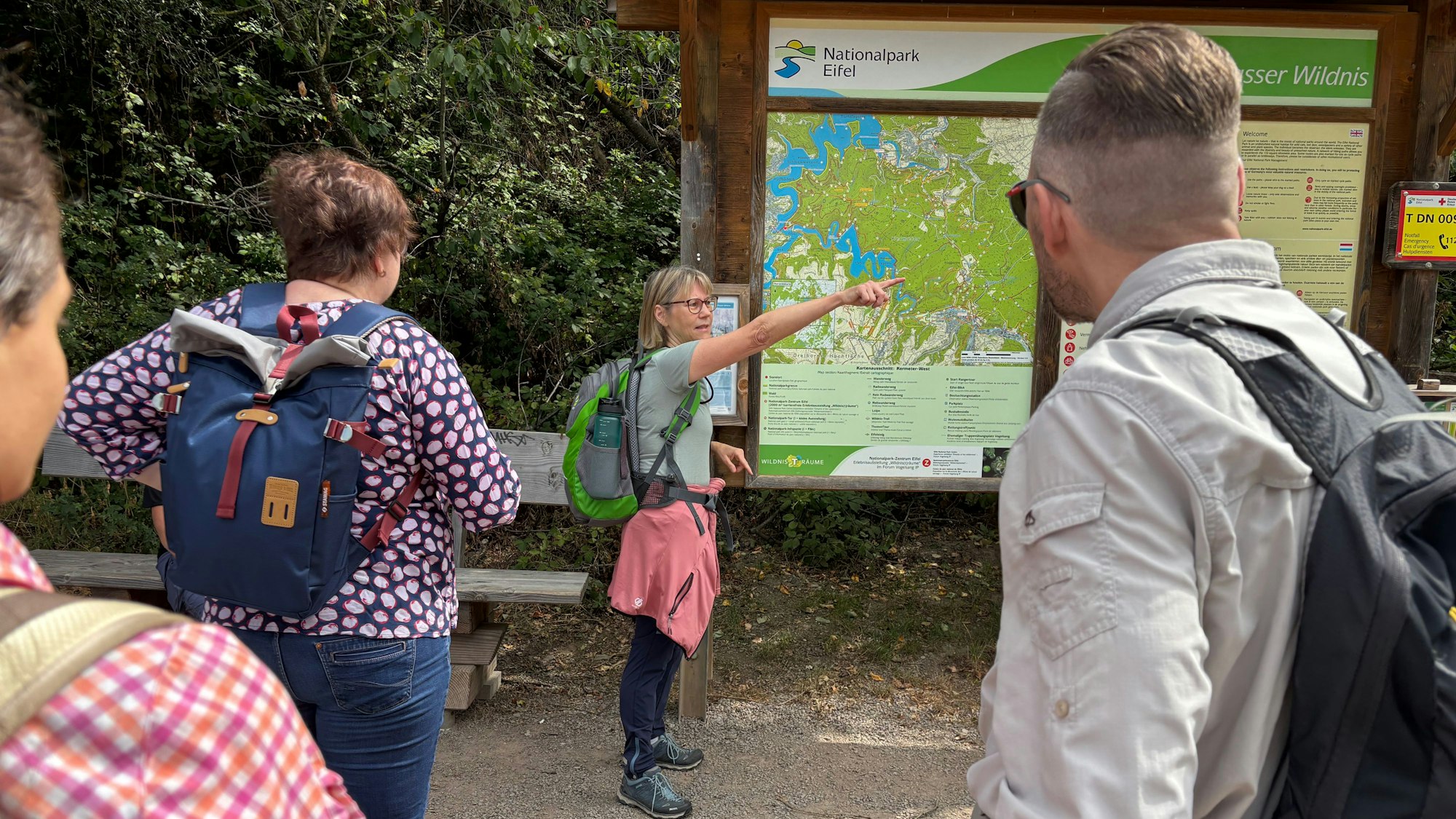Elke Griskewitz trägt einen Wanderrucksack und zeigt vor einer Karte auf etwas, das außerhalb des Bildes liegt.