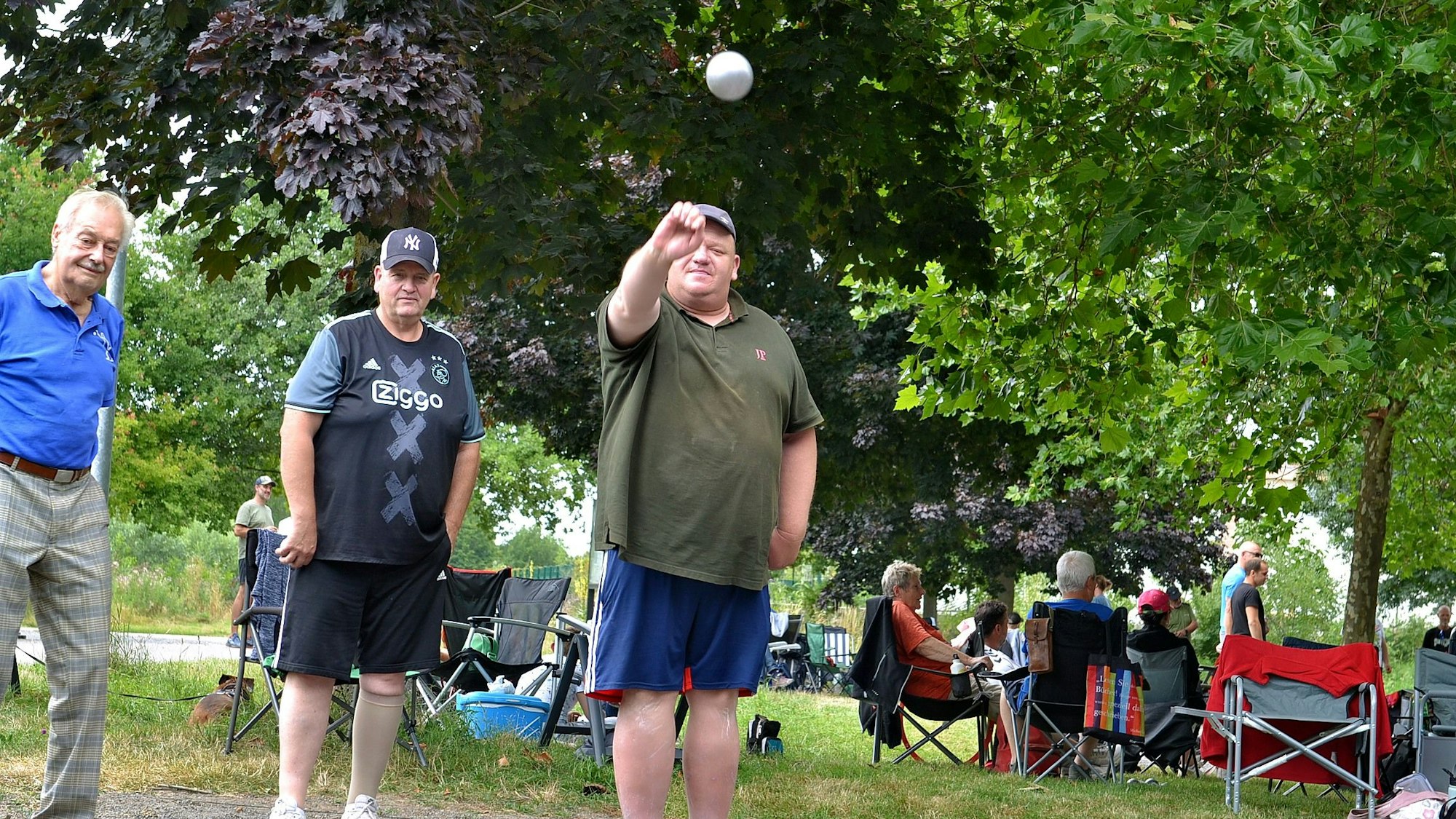Ein Mann wirft eine Boule-Kugel, die sich bei der Aufnahme in der Luft befindet. Neben ihm stehen zwei weitere Männer. Im Hintergrund sitzen läute auf Campingstühlen.