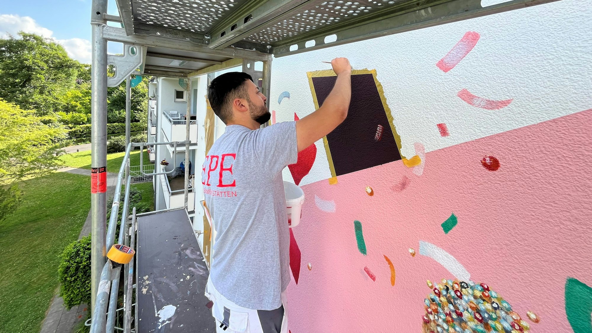 Ein Azubi der Epe Malerwerkstätten GmbH in Köln ist im Einsatz für ein Projekt gegen Vorurteile im Malerhandwerk und zeichnet an der kunstvollen Gestaltung einer Hausfassade in Porz.