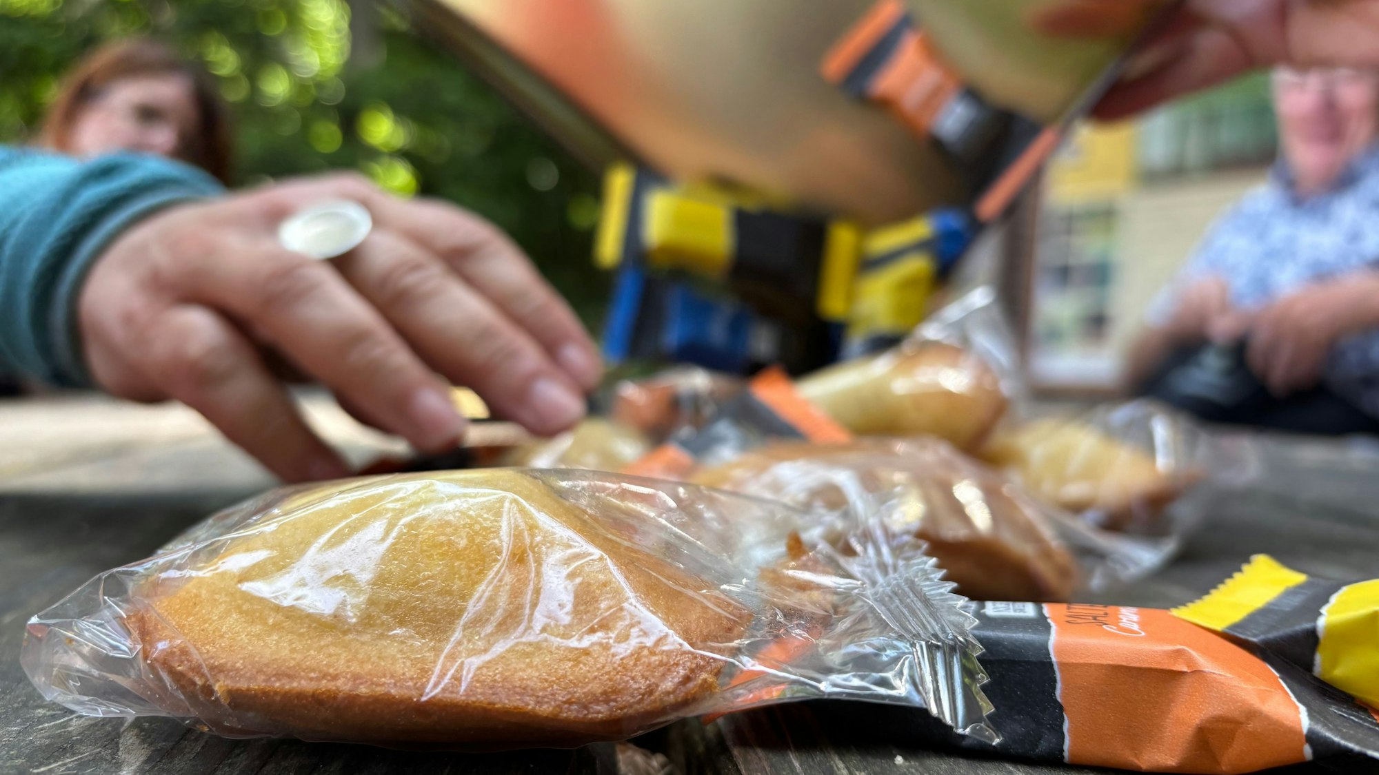 Auf einem Holztisch liegen verpackte Süßigkeiten im Vordergrund. Im unscharfen Hintergrund werden weitere Süßigkeiten ausgeschüttet und Menschen sitzen am Tisch.