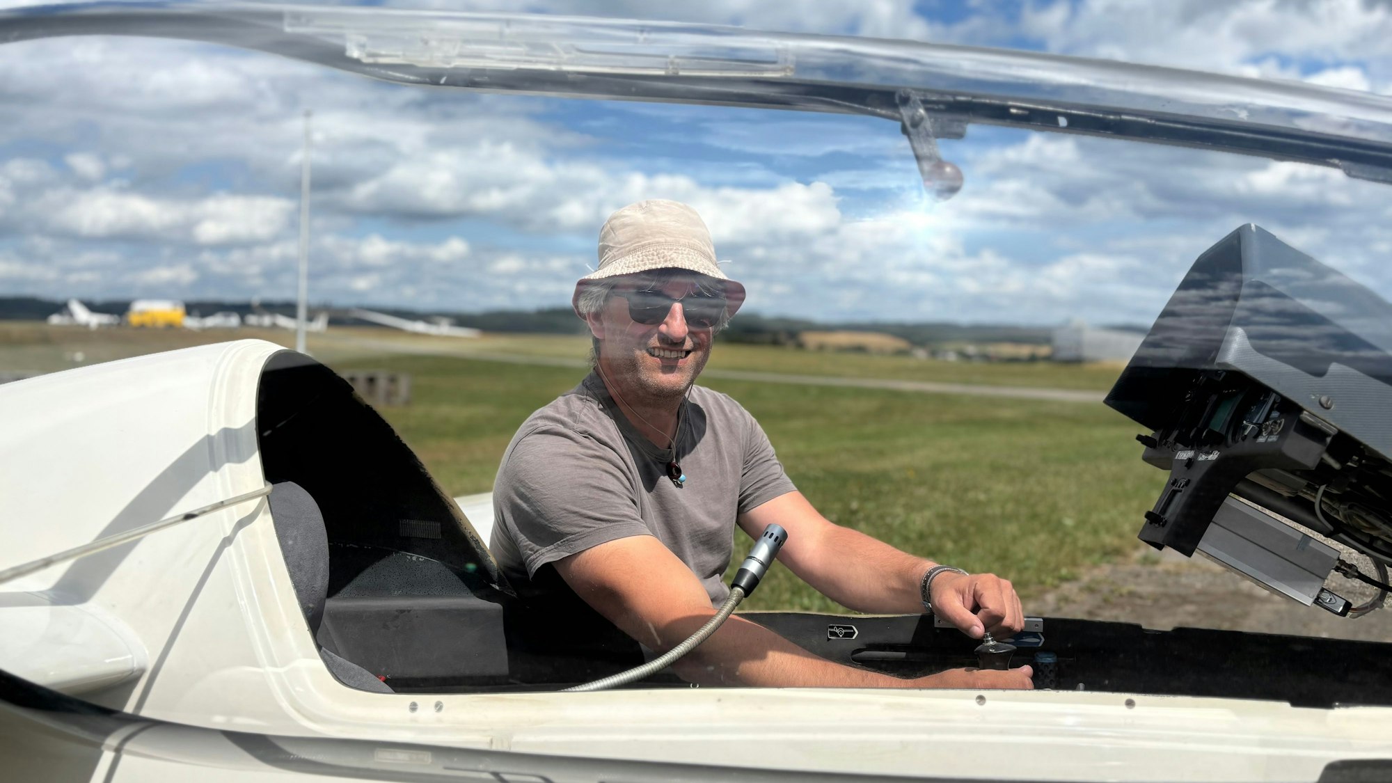 Uwe Bodenheim trägt einen Hut und eine Sonnenbrille. Er sitzt in einem Segelflugzeug mit einem Sitz, dessen Haube aufgestellt ist.