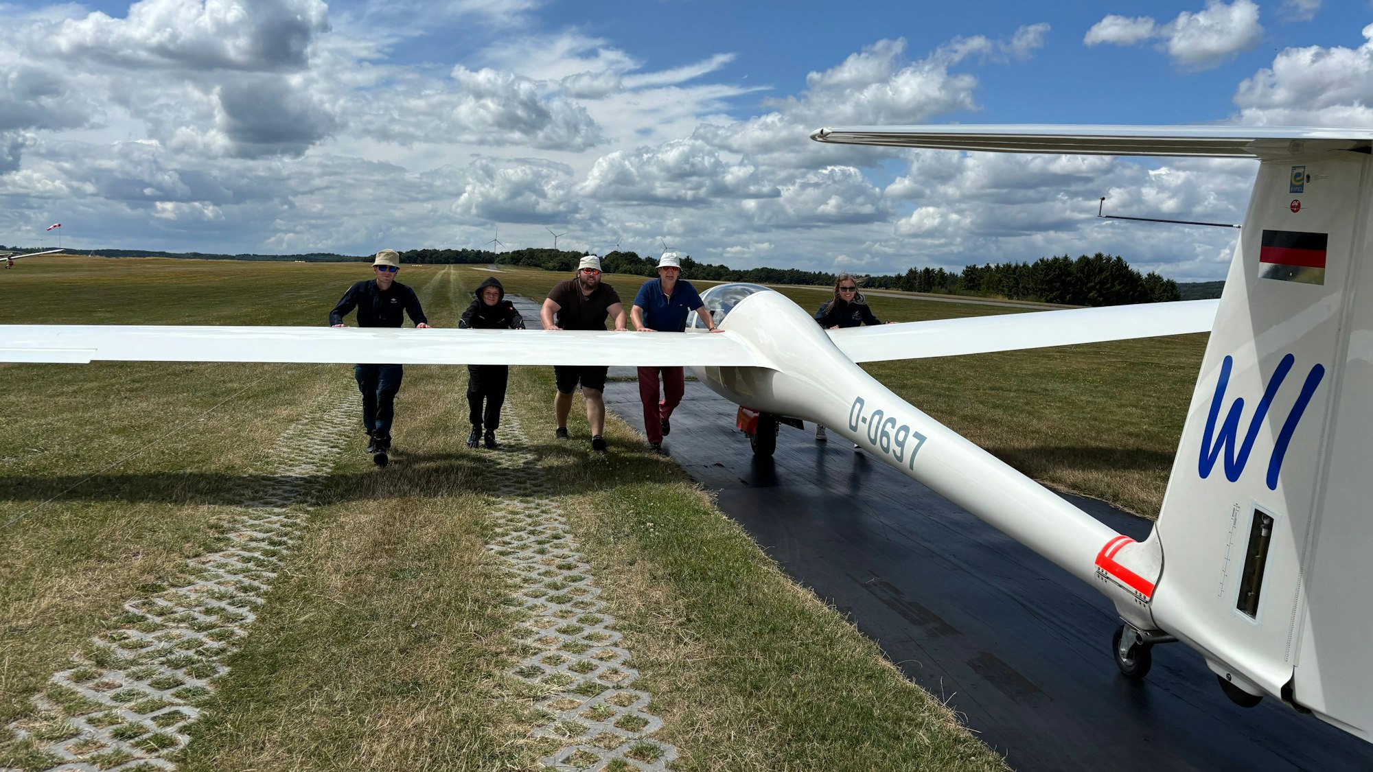 Fünf Menschen mit Sonnenbrillen und Kopfbedeckungen schieben ein Segelflugzeug über eine asphaltierte Bahn.