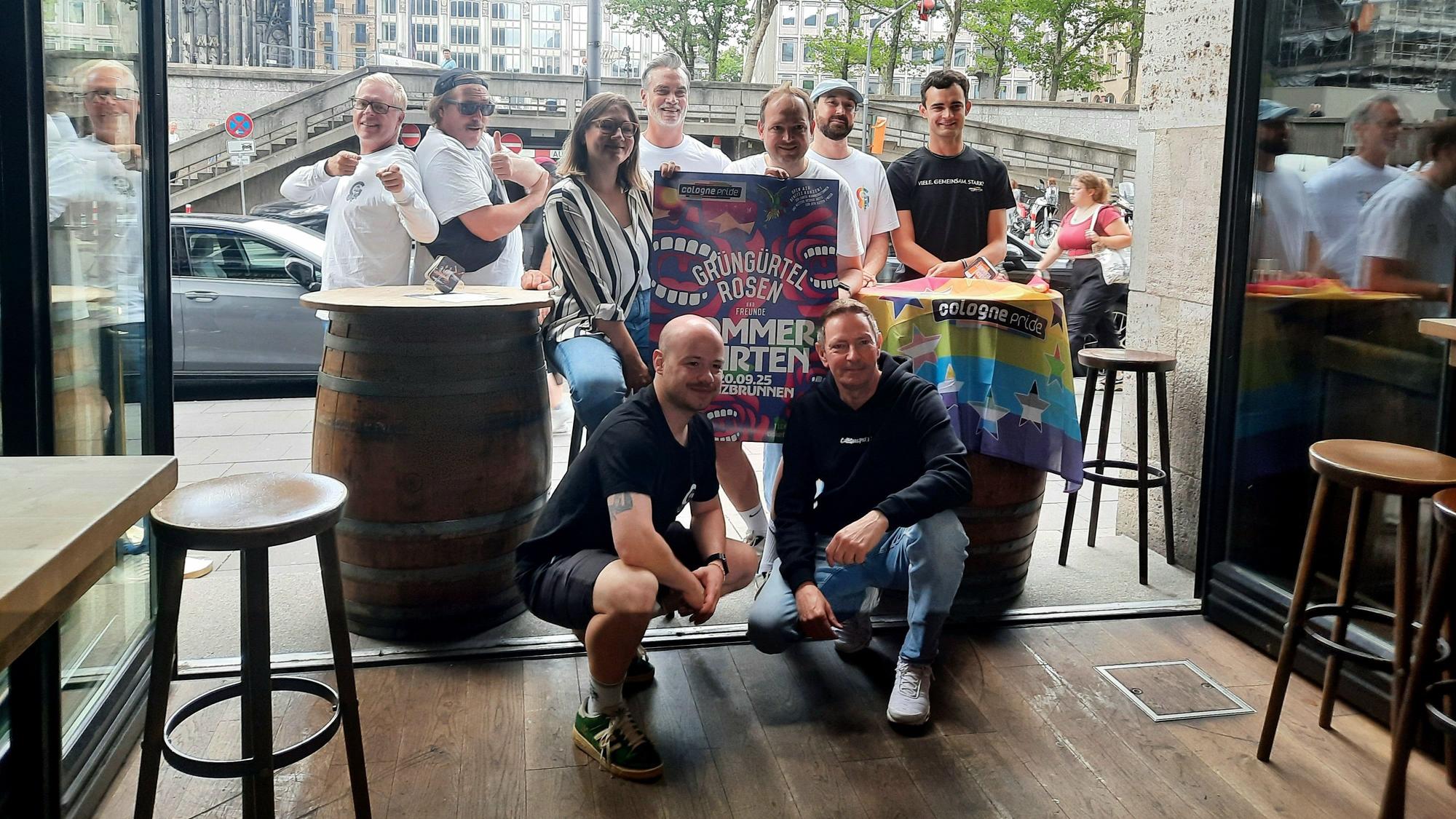 Mitglieder der „Grüngürtelrosen“ und des „ColognePride“ posieren mit dem Plakat für das Open-Air-Konzert und einer Regenbogenflagge.