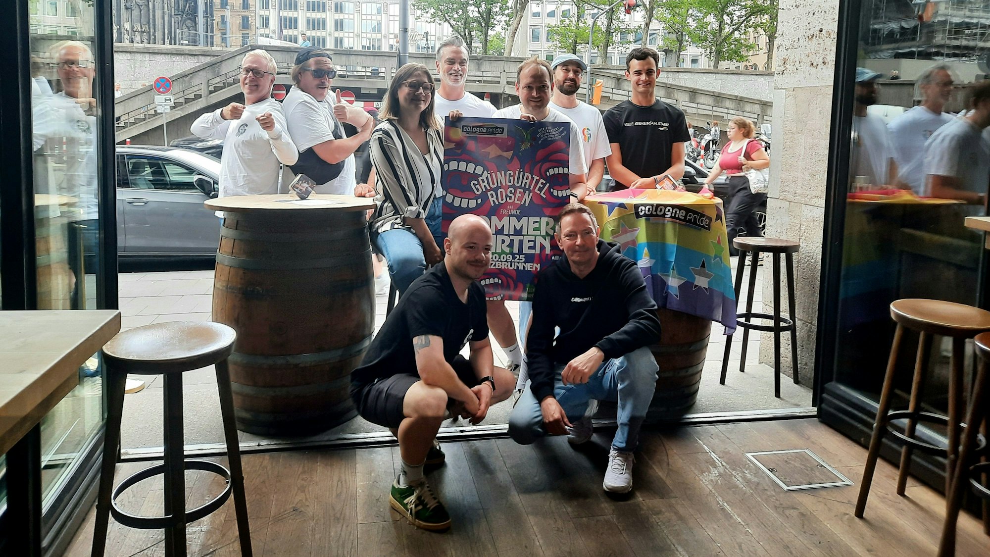 Mitglieder der „Grüngürtelrosen“ und des „ColognePride“ posieren mit dem Plakat für das Open-Air-Konzert und einer Regenbogenflagge.