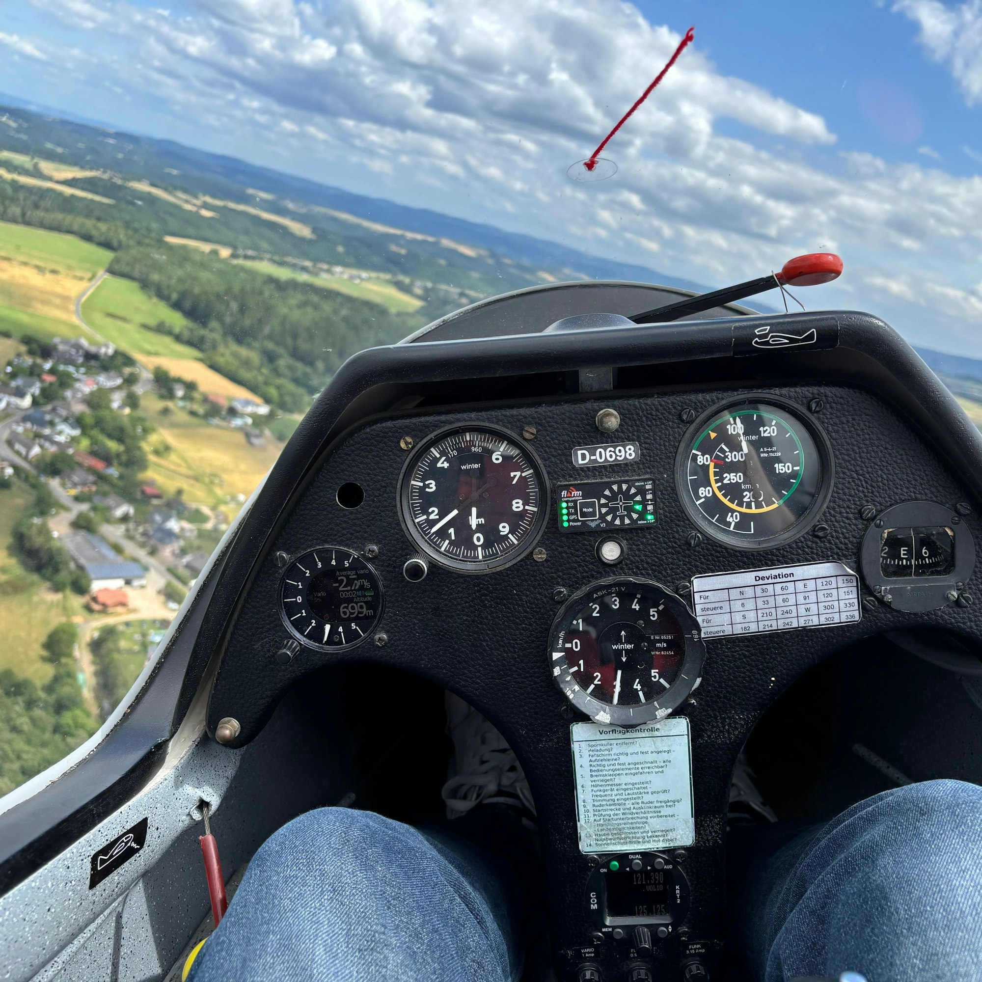 Blick aus dem vorderen Teil eines Segelflugzeugs mit zwei Sitzen: Hinter dem scharfgestellten Instrumentenbrett befinden sich Felder, Wälder und Gebäude am Boden.