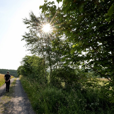 Eine Frau geht bei Sonnenschein zwischen Feldern mit ihrem Hund spazieren.