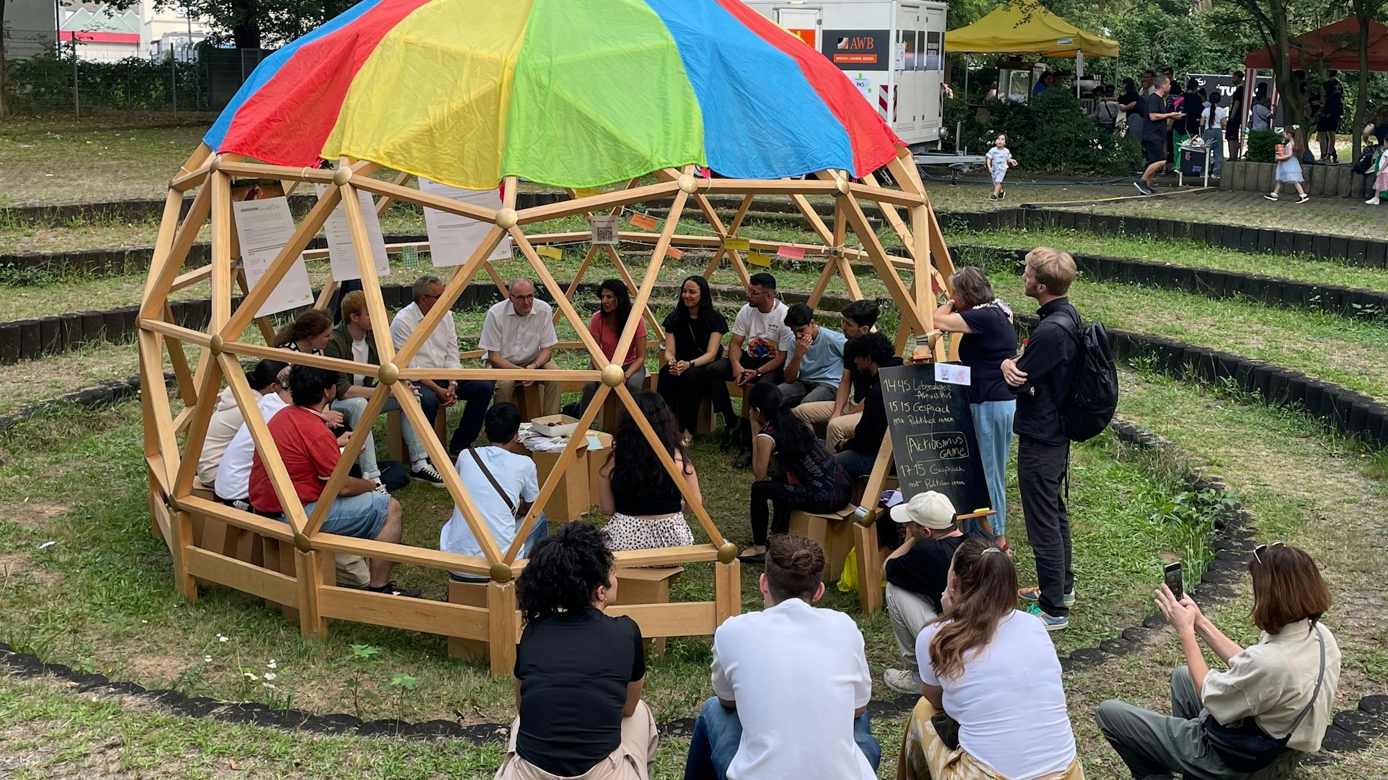 Beteiligung und Kontakt auf Augenhöhe: Jugendliche bei einer Veranstaltung vor der Europawahl 2024. Jugendliche und Politiker sitzen in einem Rundbau mit buntem Sonnenschutz zusammen und sprechen über Themen, der Jugenddpolitik.