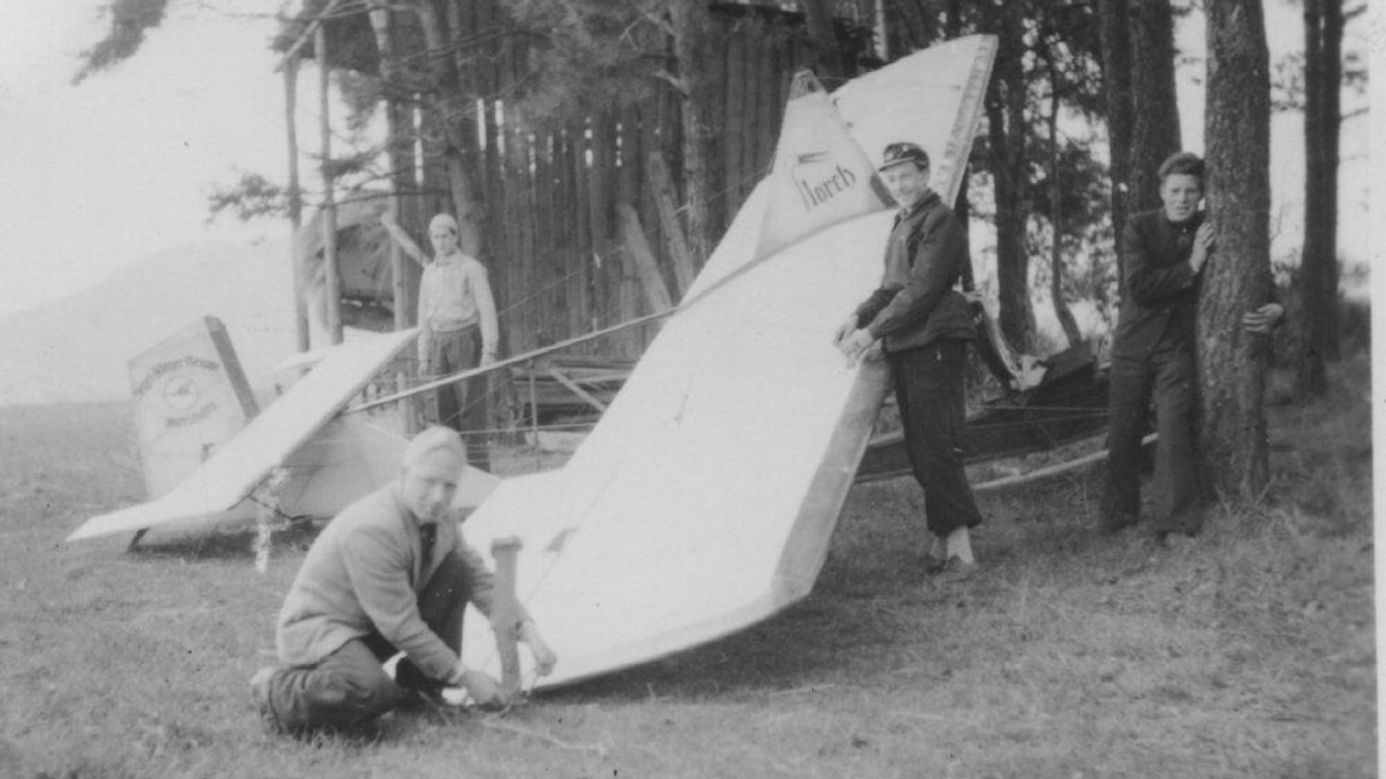 Auf einer Schwarzweiß-Aufnahme aus den 1950er Jahren stehen drei Männer und ein Jugendlicher um eine Segelflugmaschine. Diese liegt vor Bäumen auf einer Wiese.