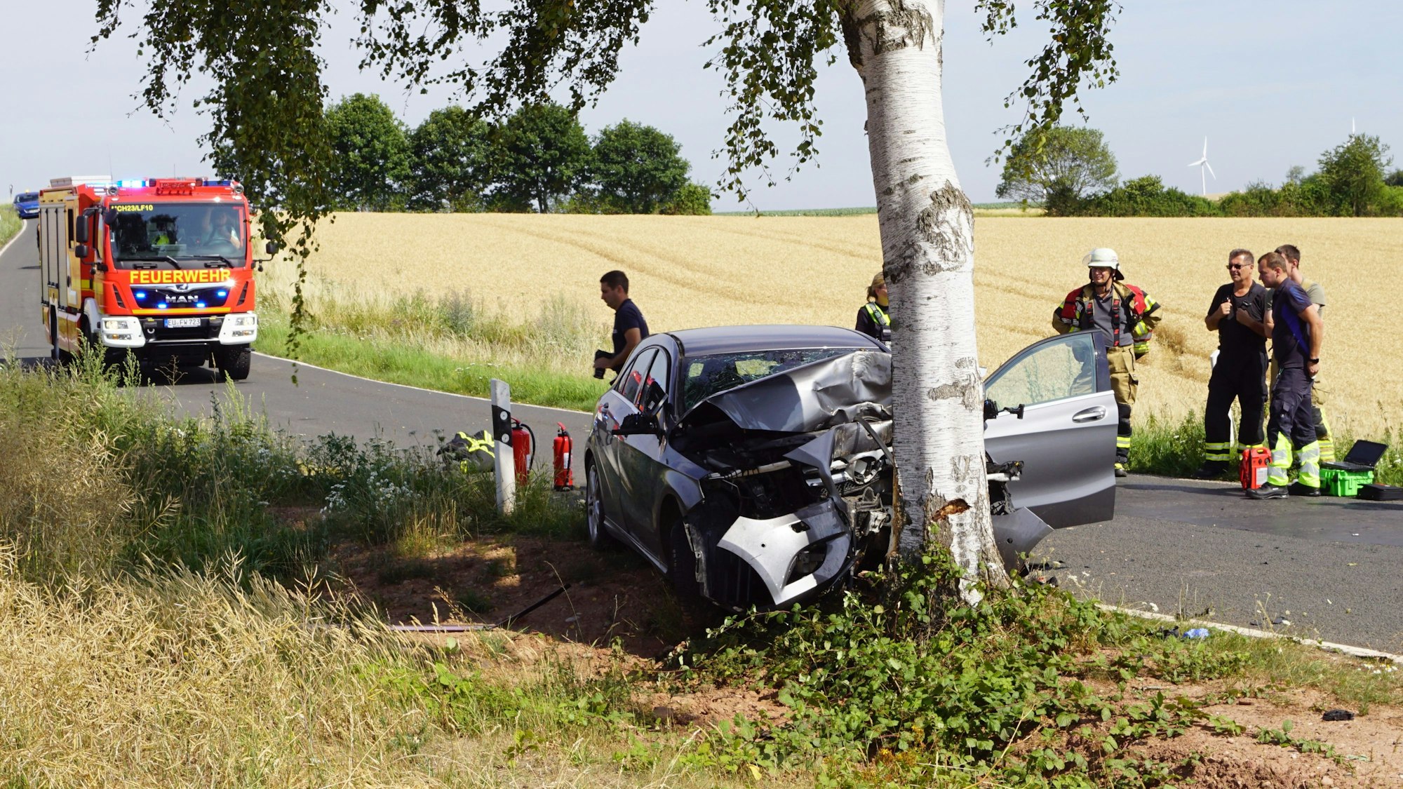 Das Foto zeigt den zerstörten Pkw am Baum, daneben Polizisten und Feuerwehrleute und im Hintergrund auf der Straße ein Feuerwehrfahrzeug.
