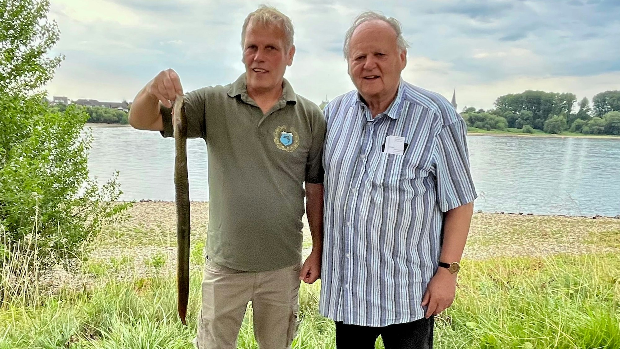Auf dem Bild sind zwei Männer am Rheinufer zu sehen. Einer hält einen geangelten Aal in der Hand.