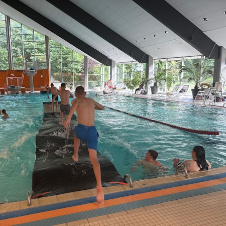 Kinder laufen in einem Hallenbad über eine Laufmatte auf dem Wasser.
