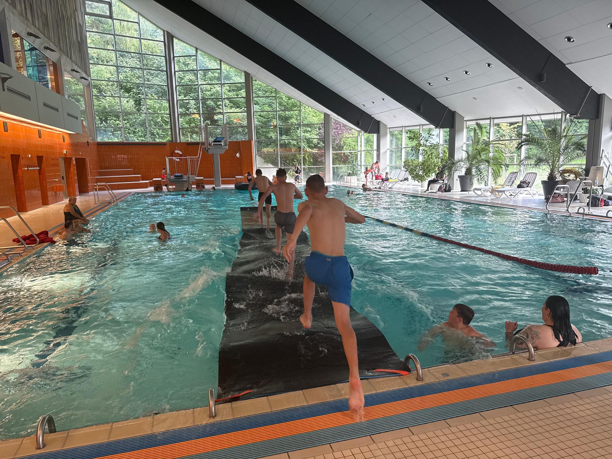 Kinder laufen in einem Hallenbad über eine Laufmatte auf dem Wasser.