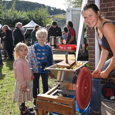Eine Frau zerkleinert Äpfel mit einer Kurbel, daneben stehen zwei Kinder.
