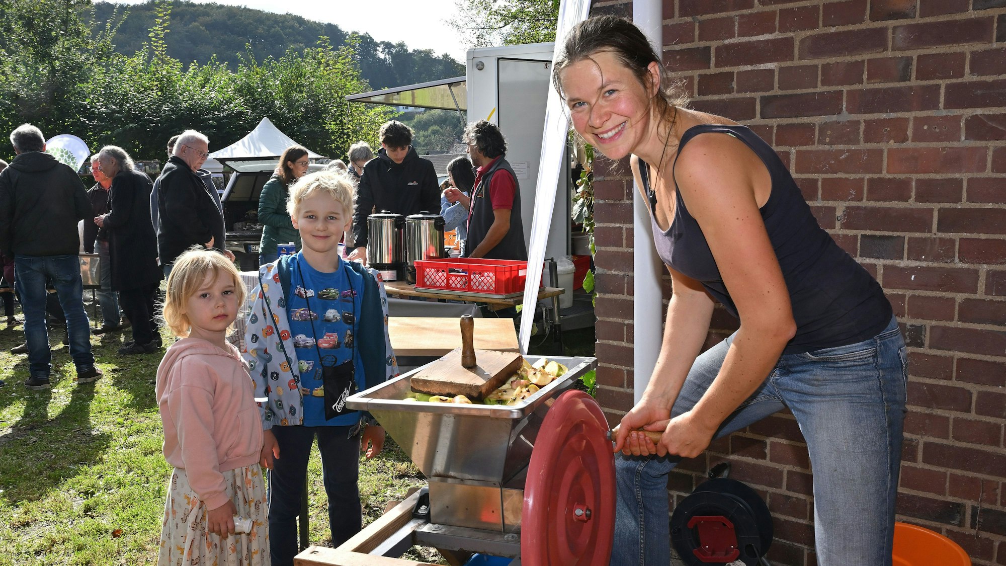 Eine Frau zerkleinert Äpfel mit einer Kurbel, daneben stehen zwei Kinder.