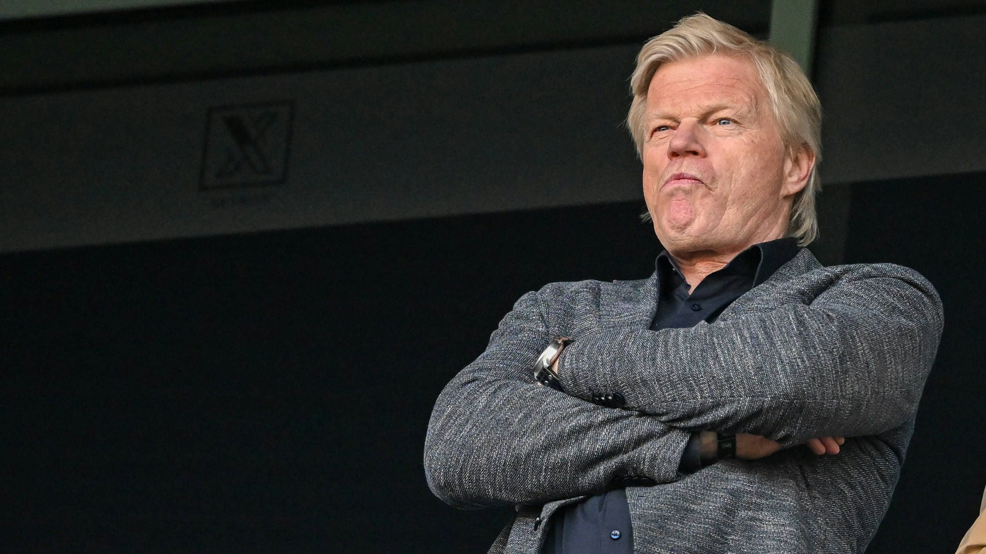 Oliver Kahn steht mit verschränkten Armen auf der Tribüne und guckt sich ein Spiel an.