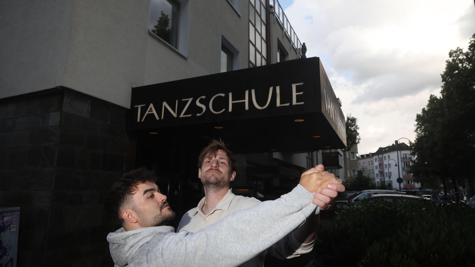 Zwei junge Männer tanzen miteinander auf offener Straße vor dem Eingang einer Tanzschule.