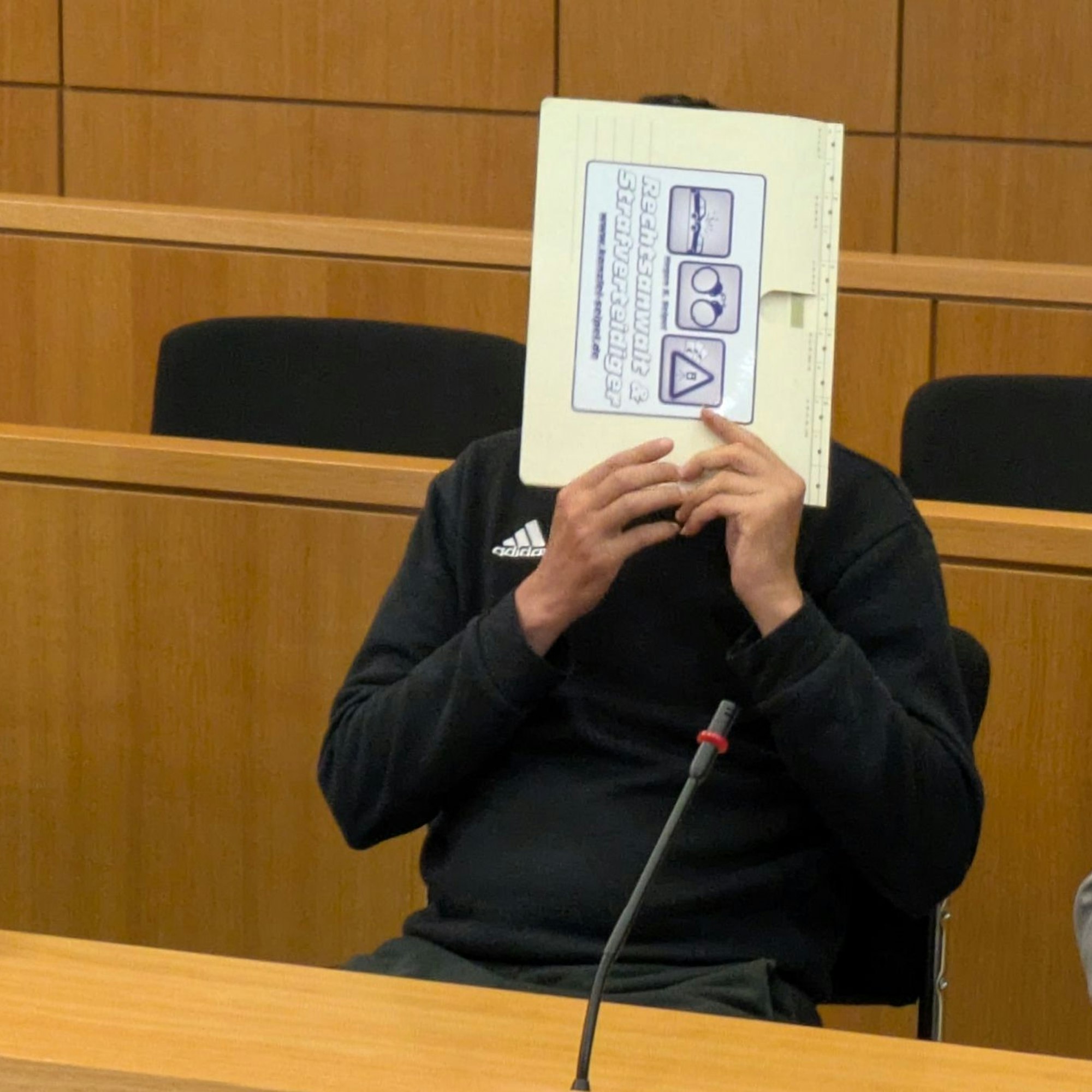 Ein Mann, der einen schwarzen Adidas-Pullover trägt, verdeckt sein Gesicht mit einer Aktenmappe.