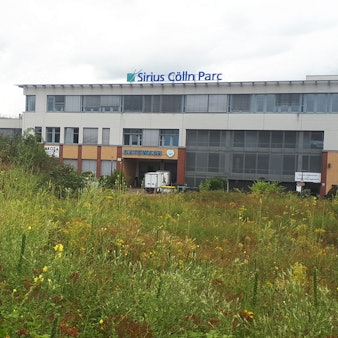 Eine grün bewucherte Brachfläche (im Vordergrund), auf der eine Dreifachturnhalle entstehen könnte. Im Hintergrund ist ein Bürogebäude von Sirius Cölln Parc zu sehen.