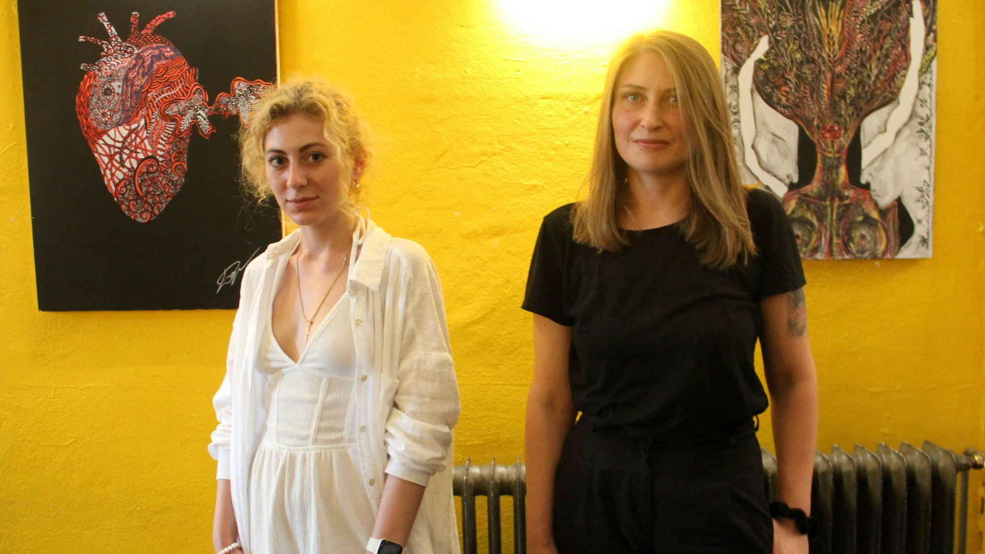 Zwei Frauen stehen vor ihren Kunstwerken, die hinter ihnen an einer gelben Wand hängen.