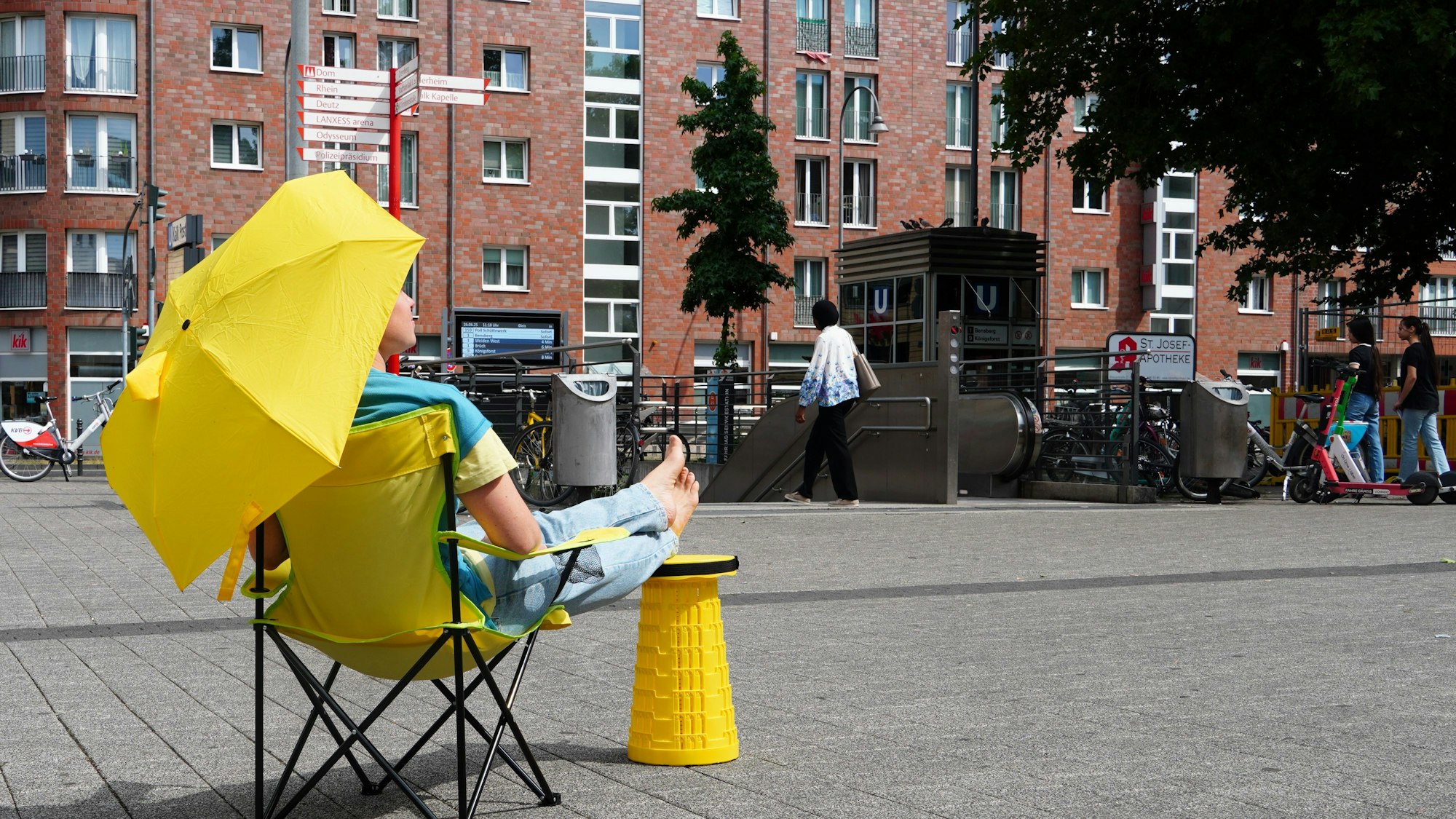 Eine Frau sitzt in einem gelben Campingstuhl, hält einen gelben Regenschirm und hat die Füße auf einem gelben Hocker hochgelegt.