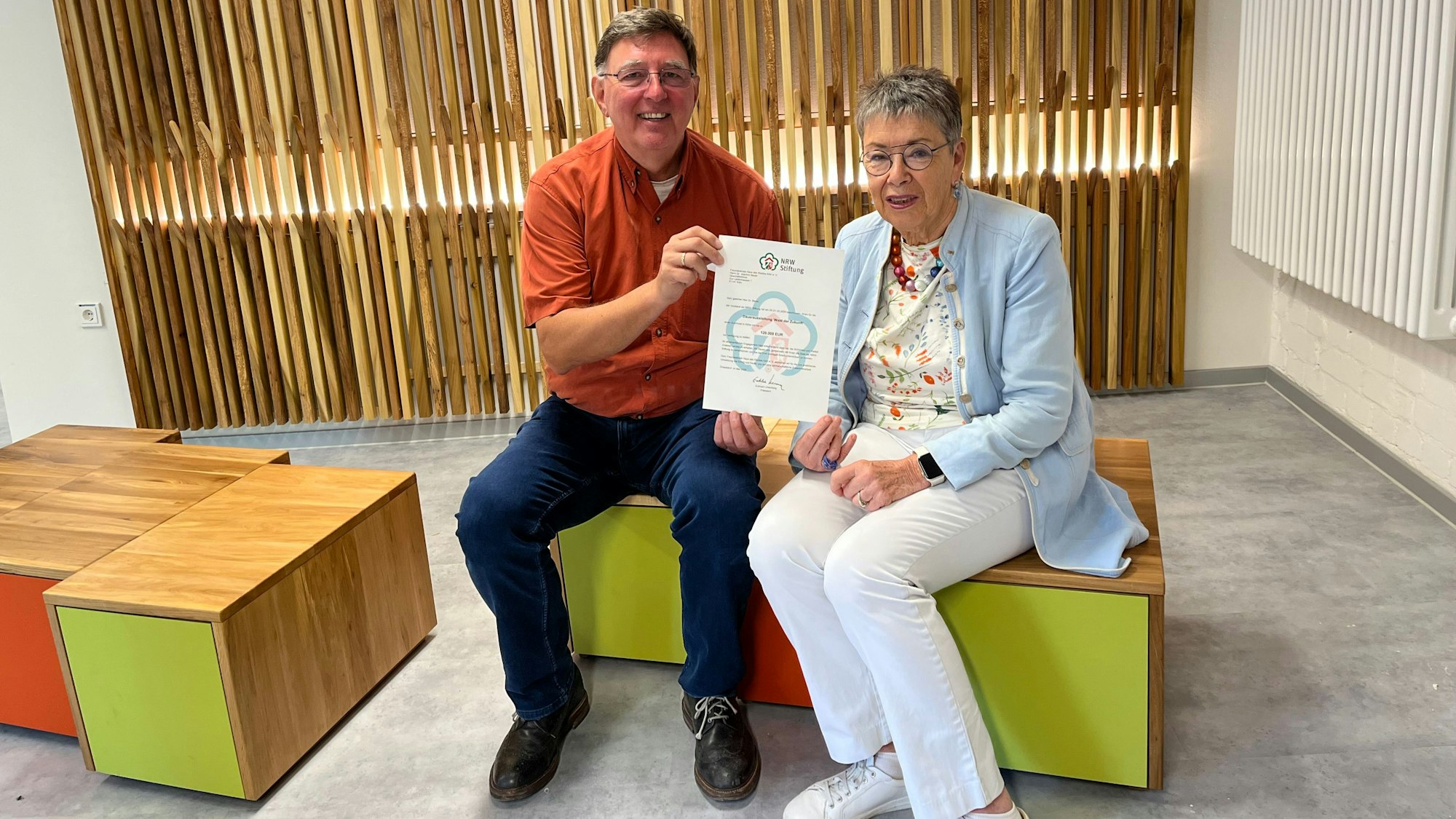 Ein Mann und eine Frau sitzen auf hölzernen Sitzwürfeln, sie halten zusammen eine Urkunde hoch.