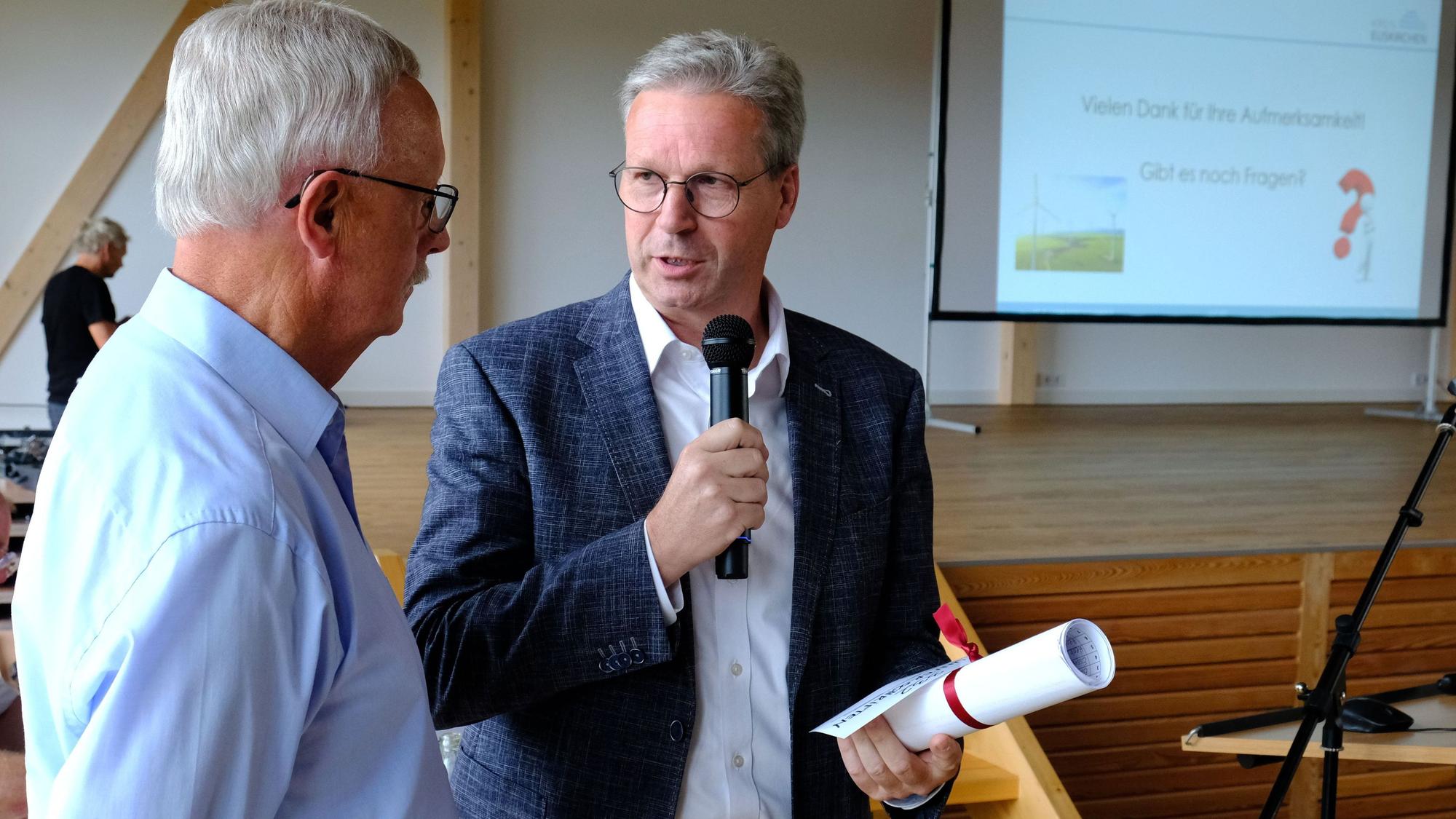 Achim Blindert (r.) spricht über ein Mikrofon mit Hubert Braun, der ihm eine zusammengerollte Unterschriftenliste überreicht hat.