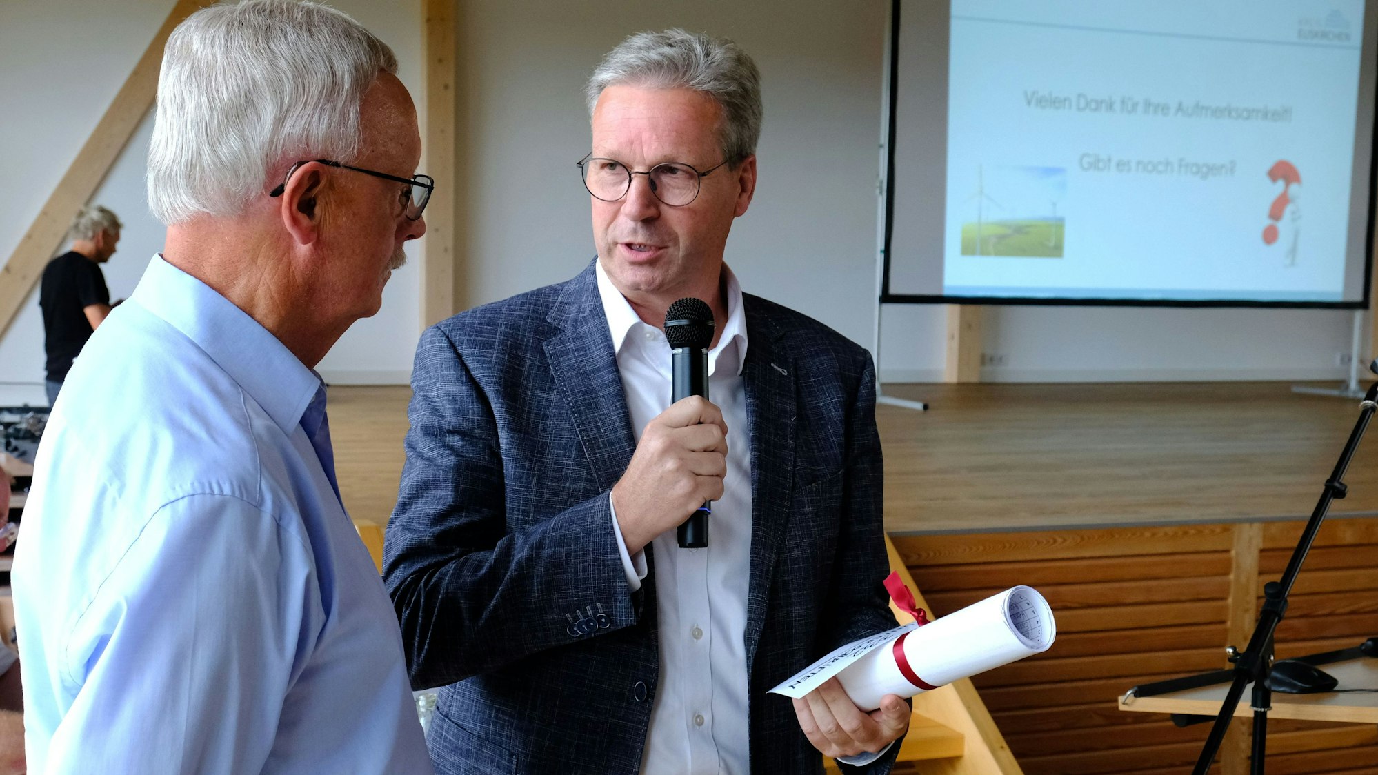 Achim Blindert (r.) spricht über ein Mikrofon mit Hubert Braun, der ihm eine zusammengerollte Unterschriftenliste überreicht hat.
