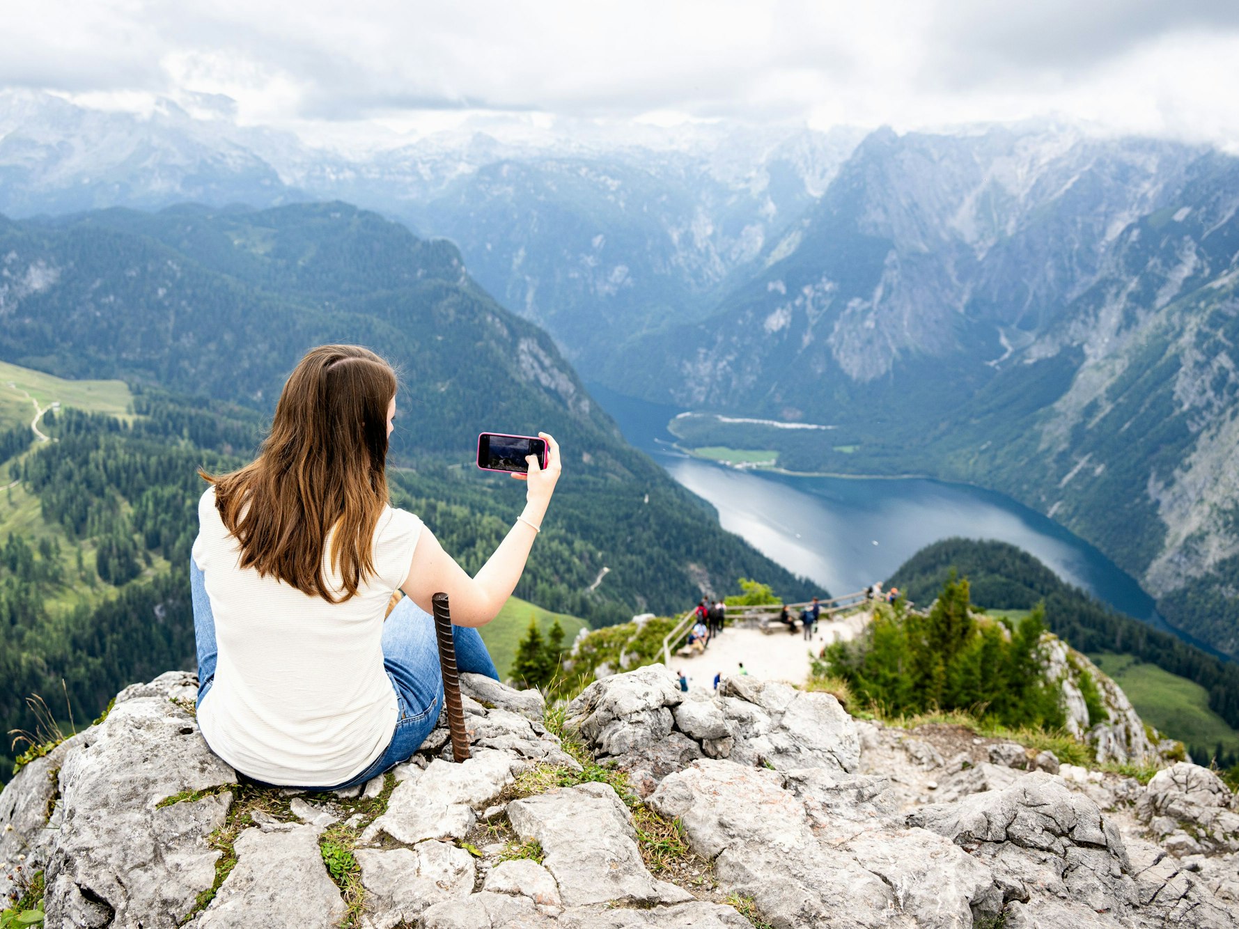 Das Bild zeigt eine junge Frau auf einem Felsen, die einen See mit dem Handy fotografiert.