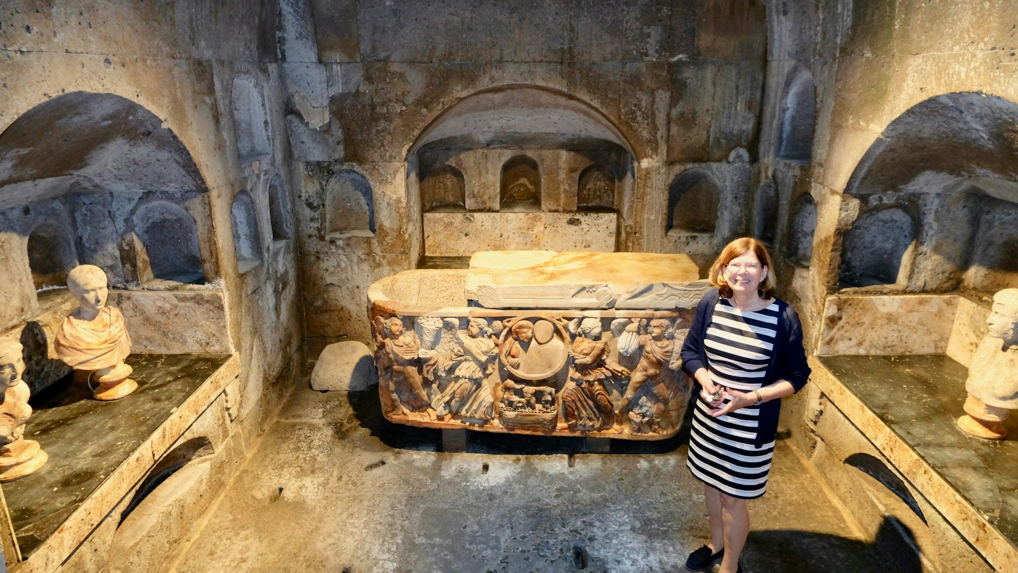 Eine Frau im Ringelkleid und Strickjacke steht inmitten einer römischen Grabkammer.