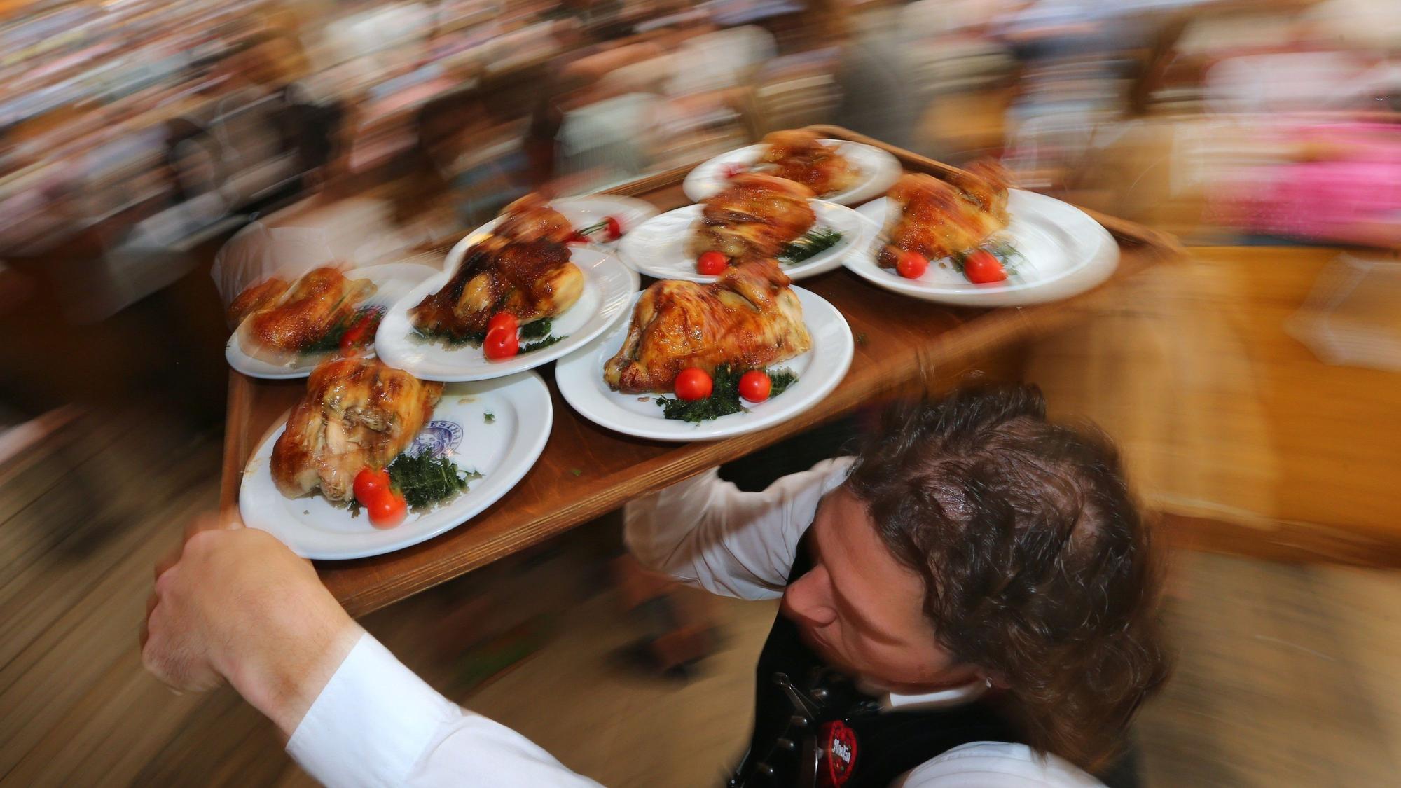 Ein Kellner trägt auf dem Oktoberfest Hendl (Hähnchen) auf einem Tablett durch ein Bierzelt.