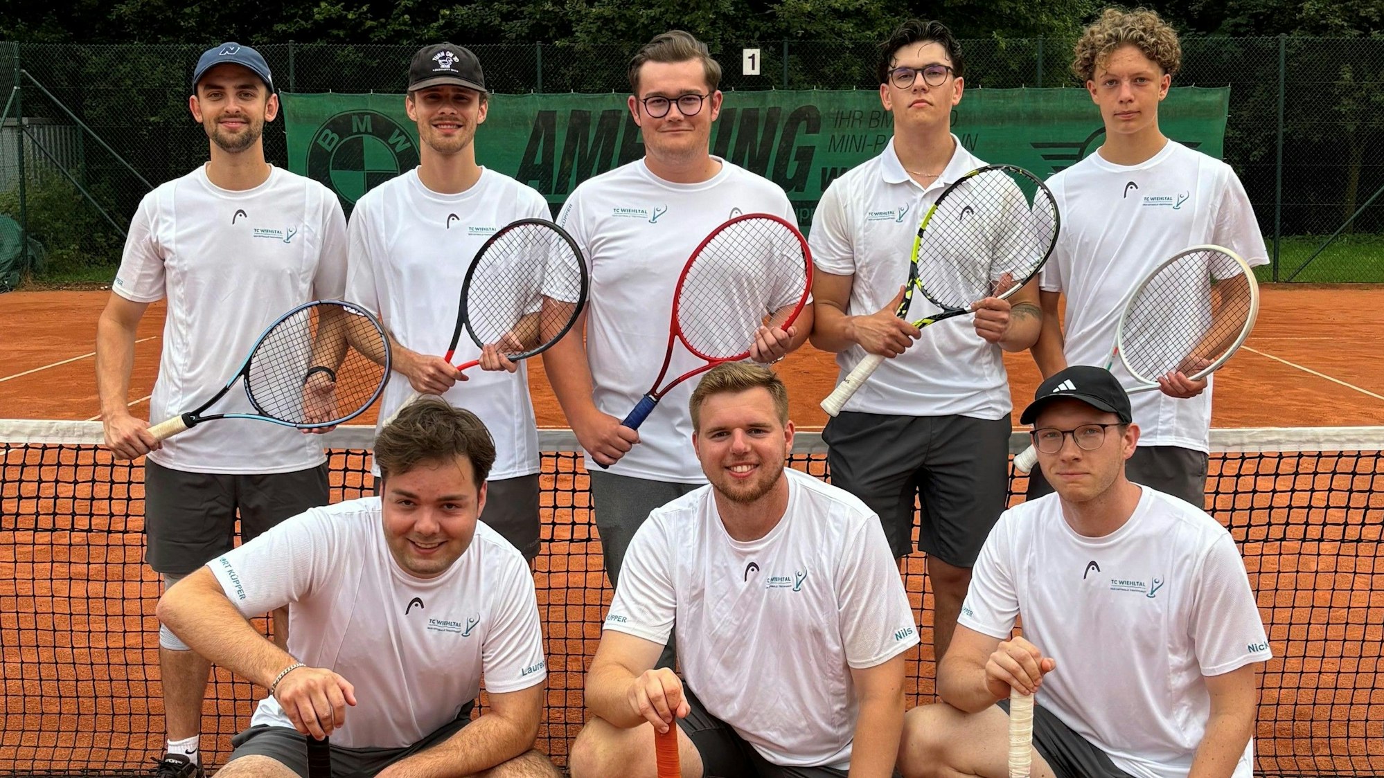 Fünf junge Männer stehen vor einem Tennisnetz mit ihren Tennisschlägern. Davor knien drei weitere junge Männer mit ihren Tennisschlägern.