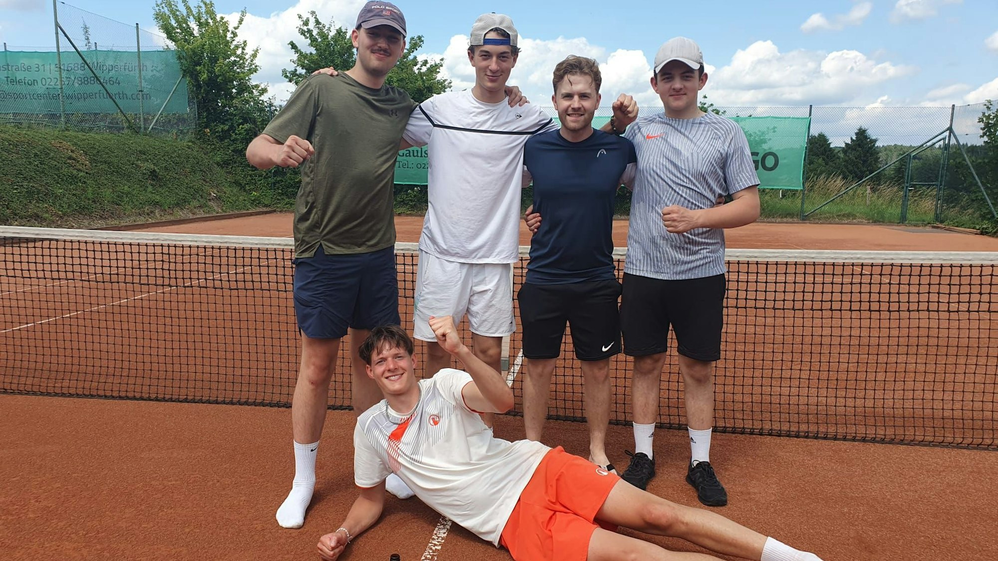 Vier junge Männer stehen vor einem Tennisnetz. Ein junger Mann liegt jubelnd davor.